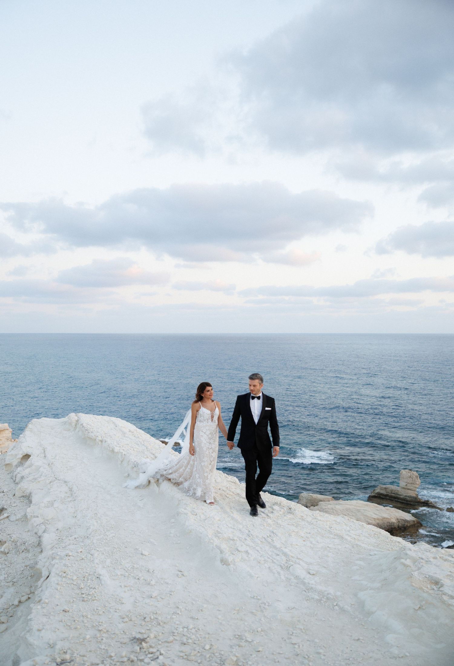 Bride and groom hold hands on white cliffside overlooking the ocean during their wedding photo session.