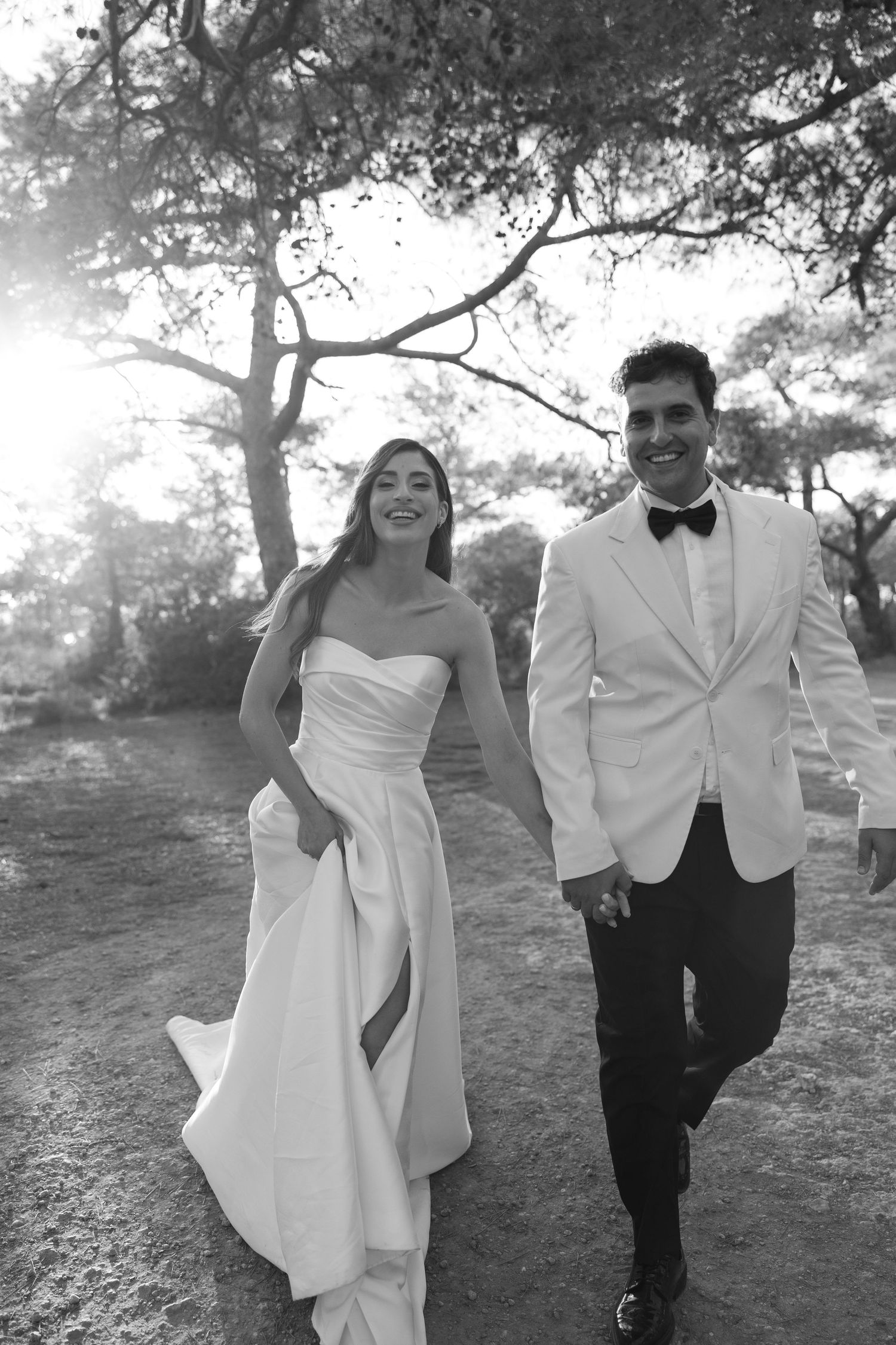 Black and white photo of a smiling bride in strapless gown and groom in white tuxedo walking hand in hand outdoors.