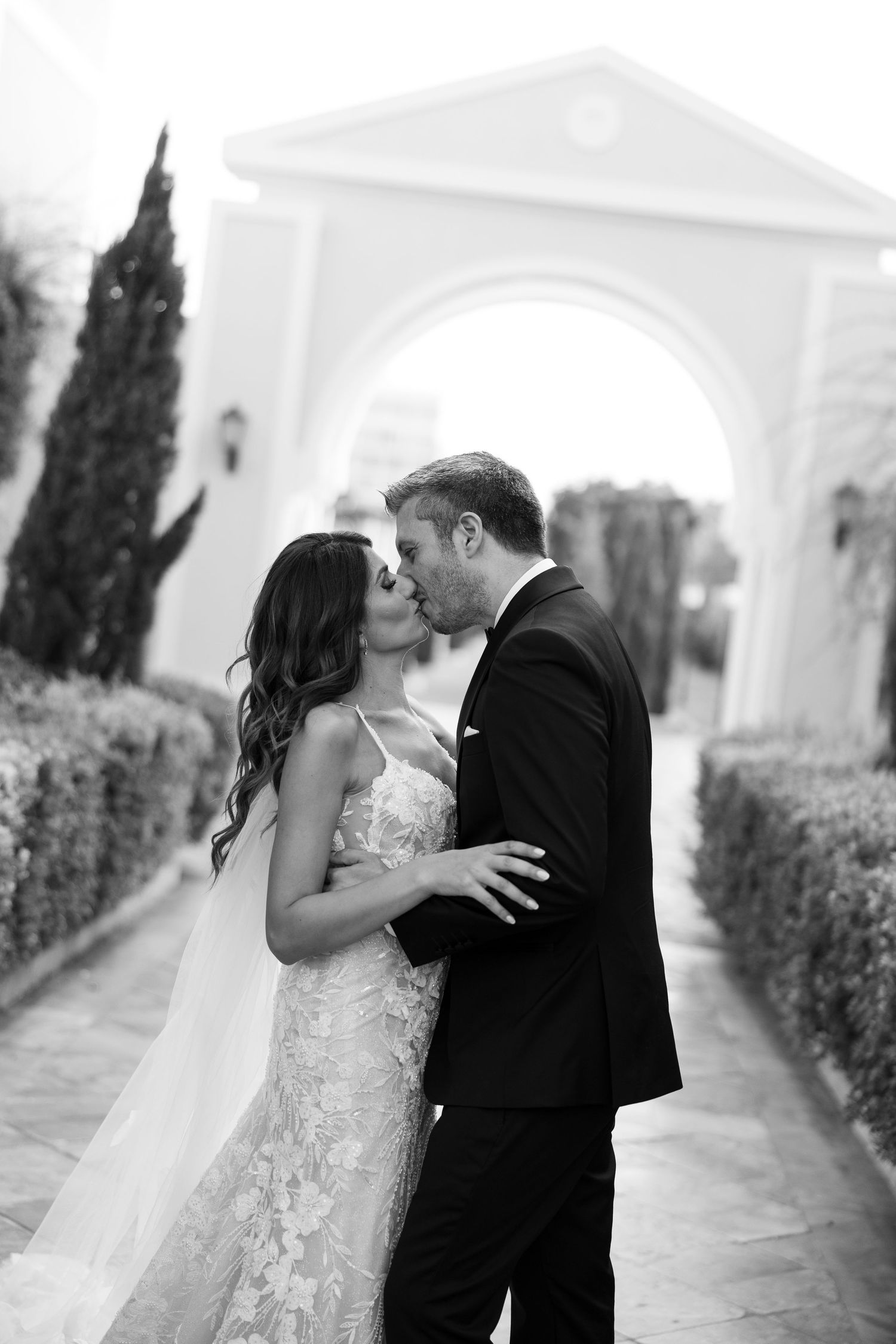 Black and white photo of bride and groom sharing a romantic kiss at elegant venue with arched entrance.