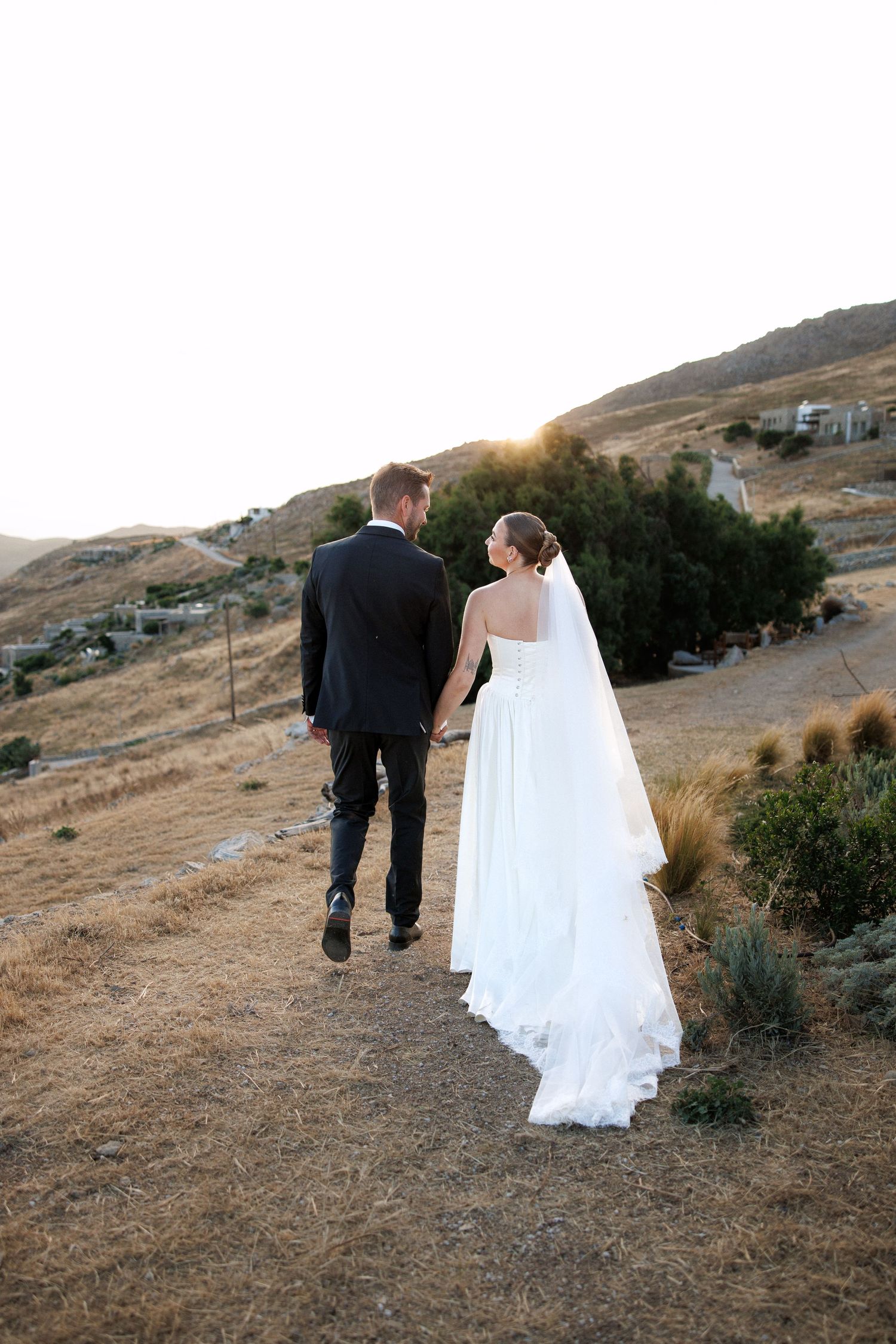 Bride and groom walking hand in hand on hillside path at sunset in wedding attire with veil trailing.