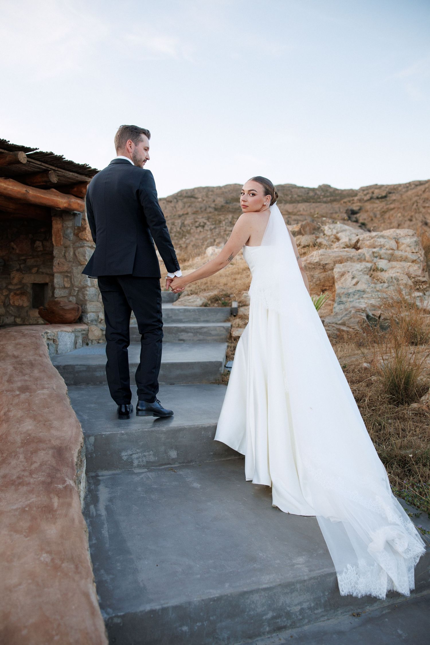 Bride in flowing white gown and veil holds grooms hand on stone steps at rustic outdoor venue.