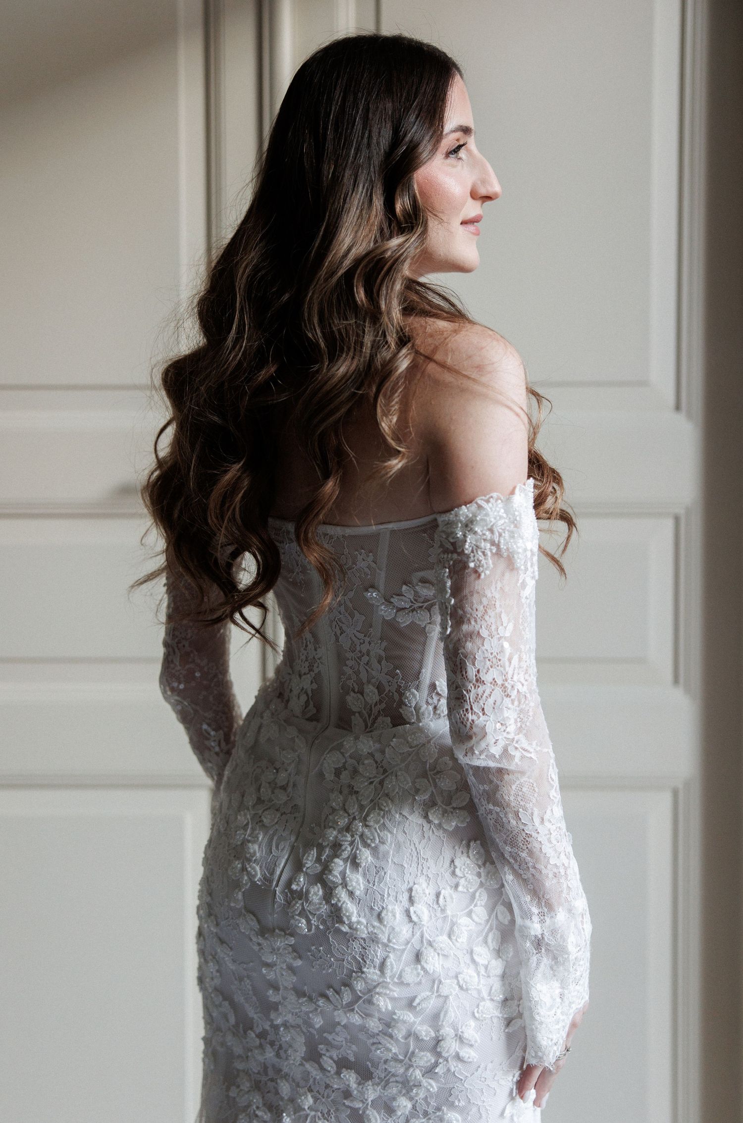 Bride in off-shoulder lace wedding gown with long sleeves and flowing dark hair, posed elegantly against a neutral wall.