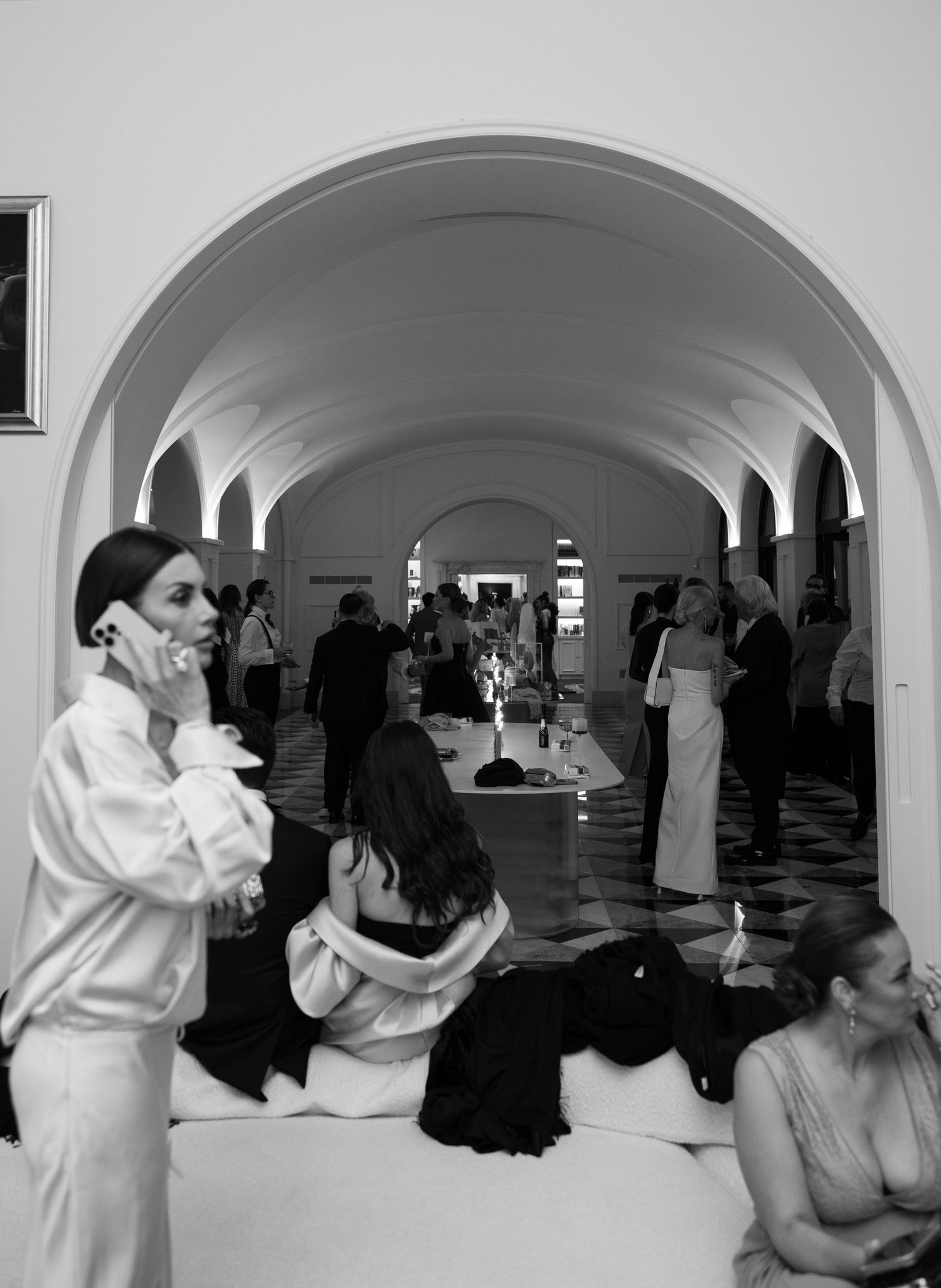 Black and white photo of elegantly dressed guests socializing in an arched hallway at a formal event.