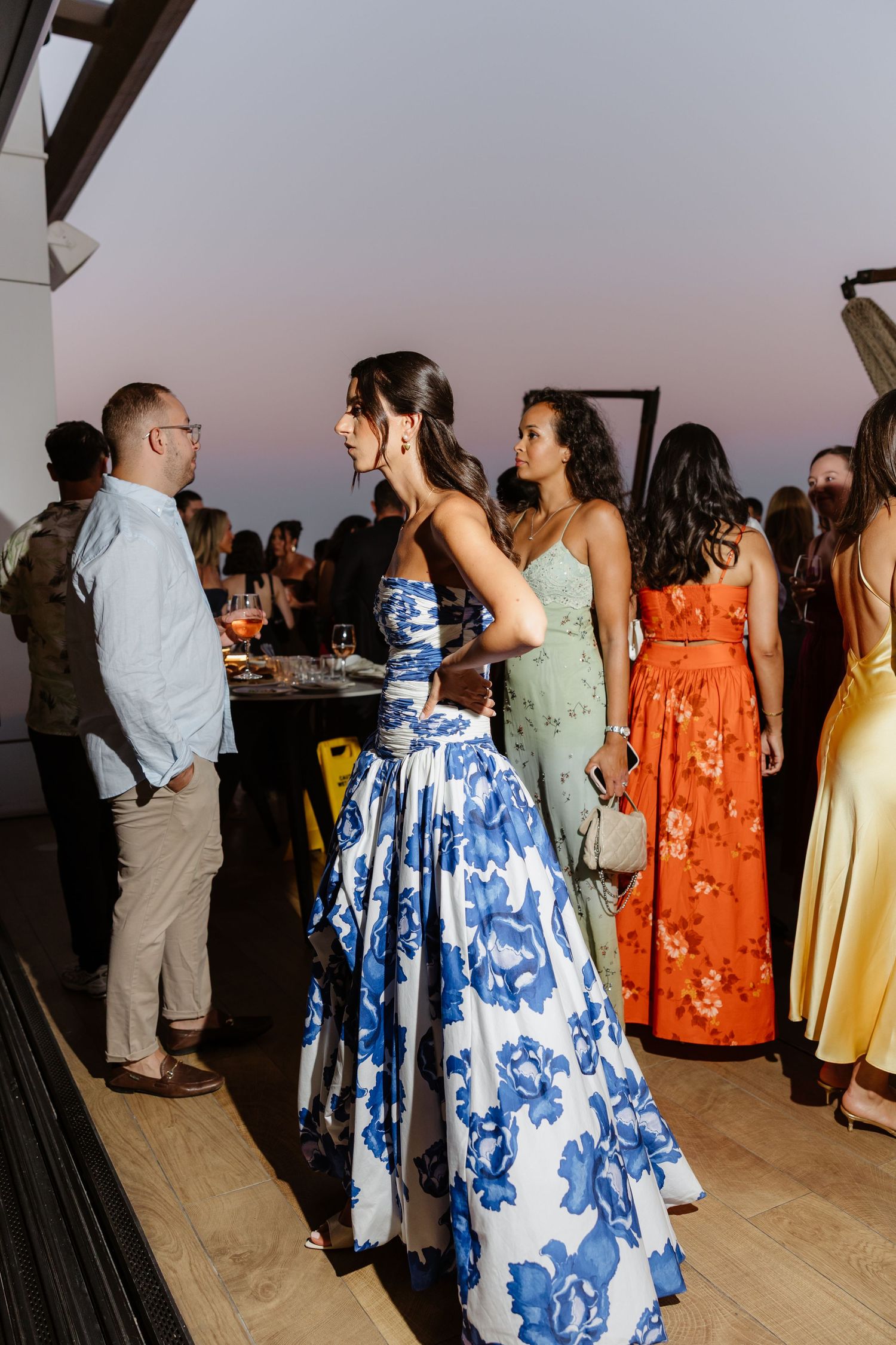 A social gathering shows attendees in colorful summer dresses including blue floral and orange designs at an outdoor venue.