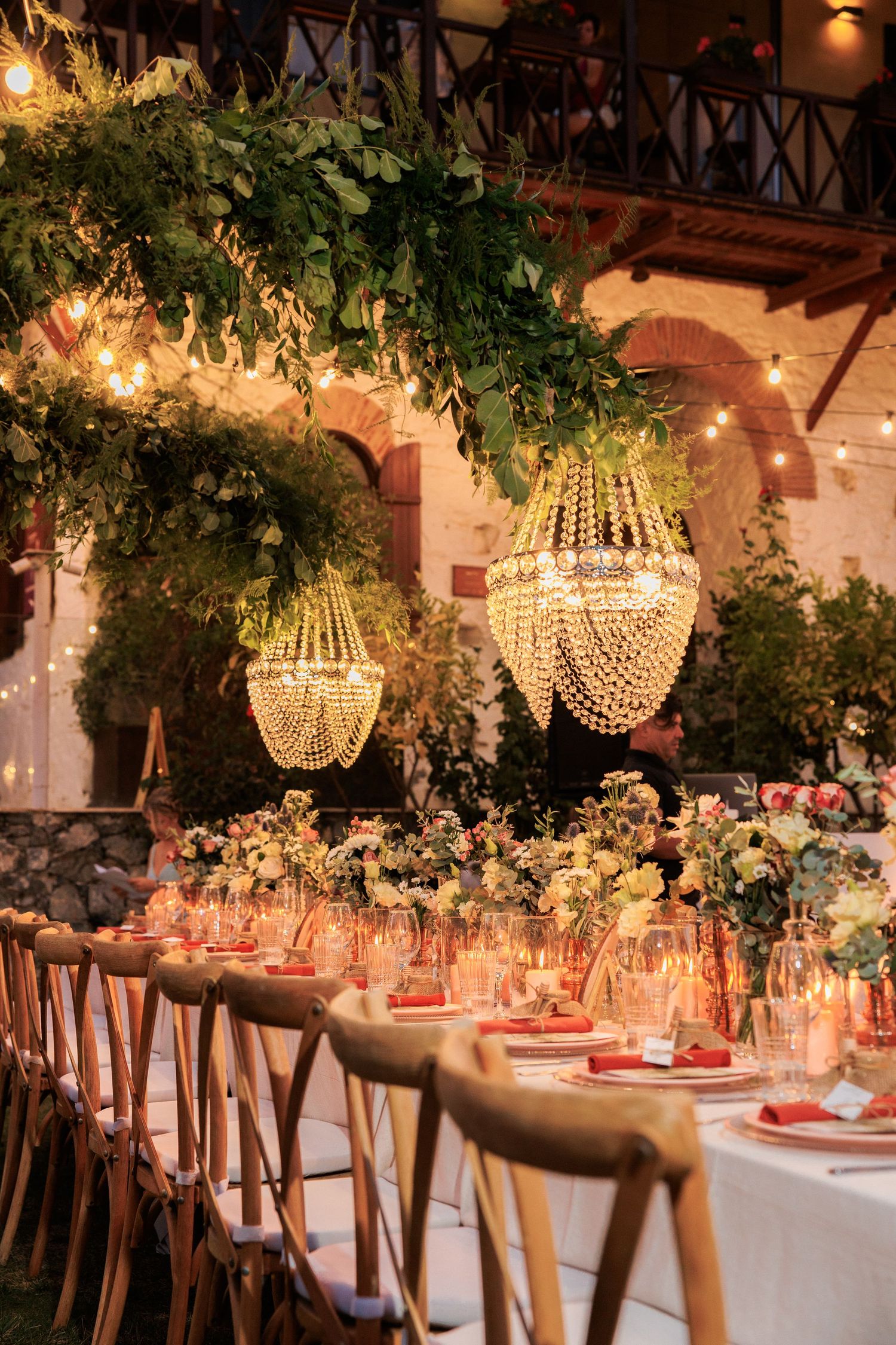 Romantic dinner table setting with crystal chandeliers and greenery under rustic pergola.
