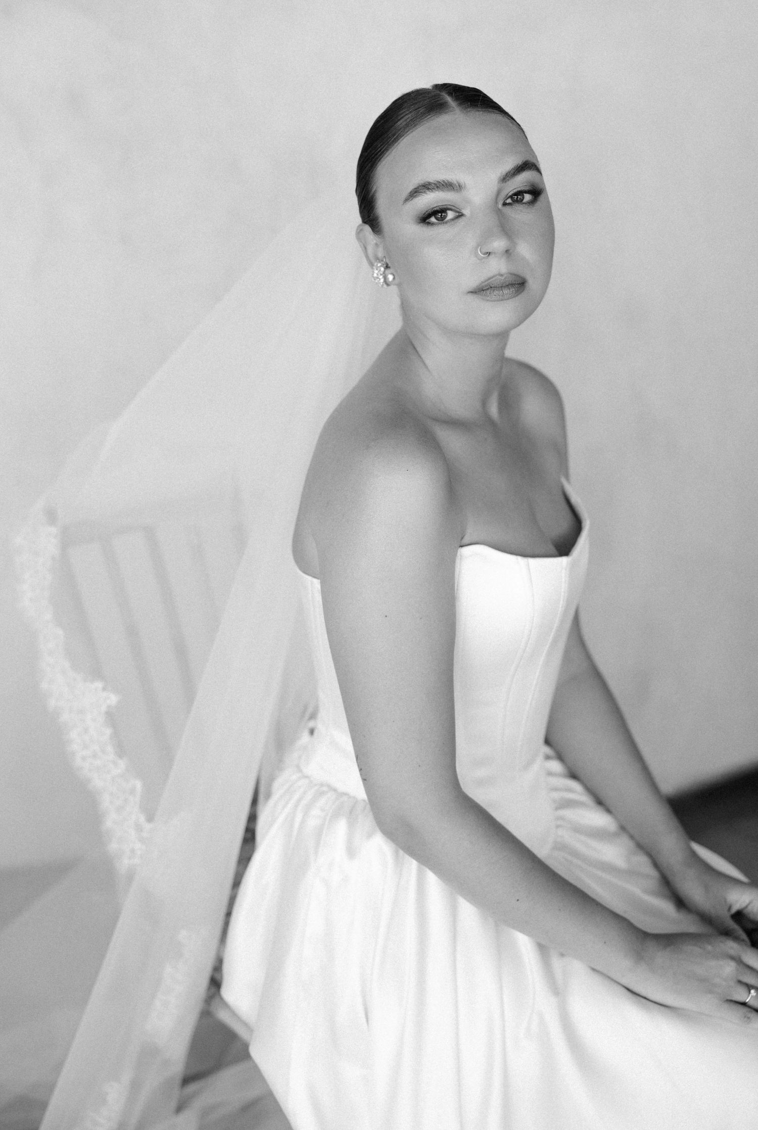 Black and white bridal portrait of a woman in strapless gown and lace-trimmed veil, seated elegantly.