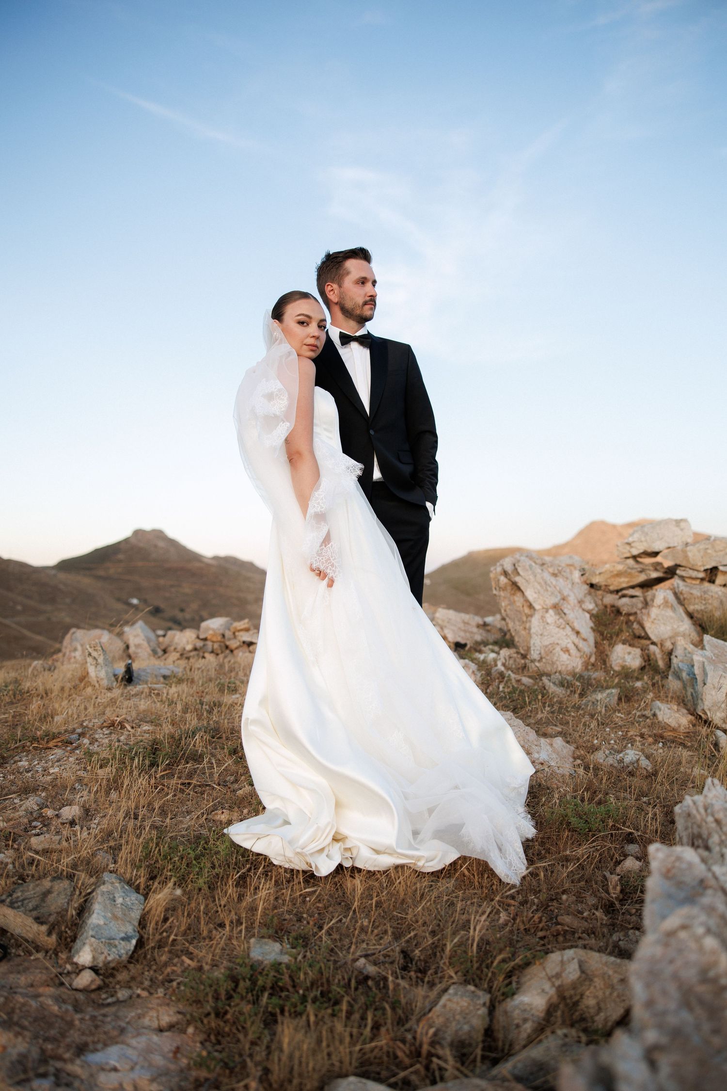Bride in flowing white gown and veil embraces groom in black tuxedo against scenic mountain landscape at sunset.