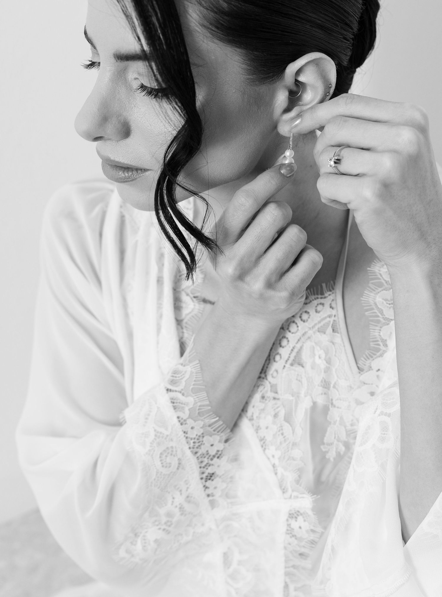 Black and white photo of a bride in lace robe putting on pearl earrings, wearing an engagement ring.