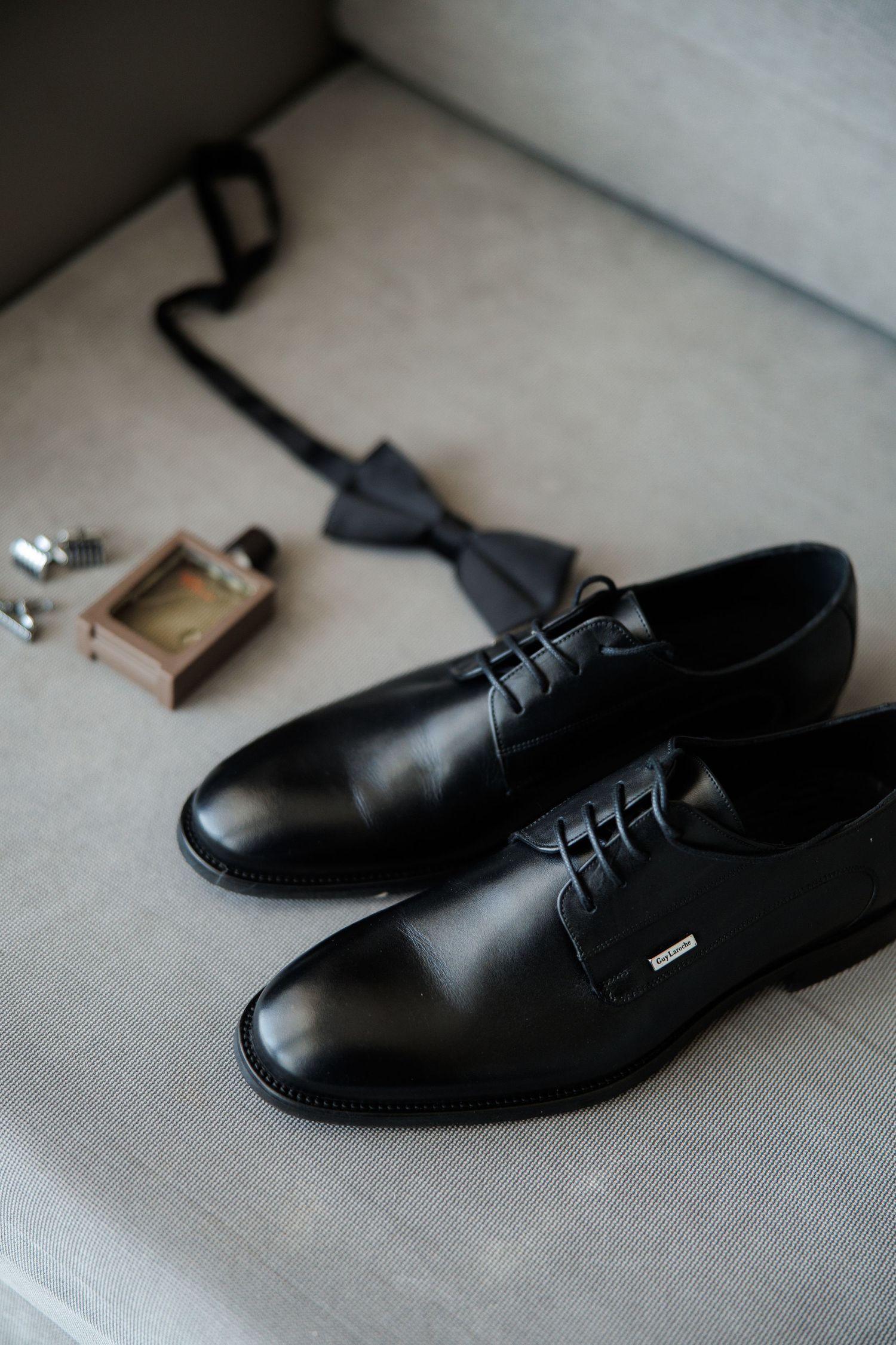Black leather oxford dress shoes with bow tie, cologne, and cufflinks arranged on a gray sofa for a wedding.