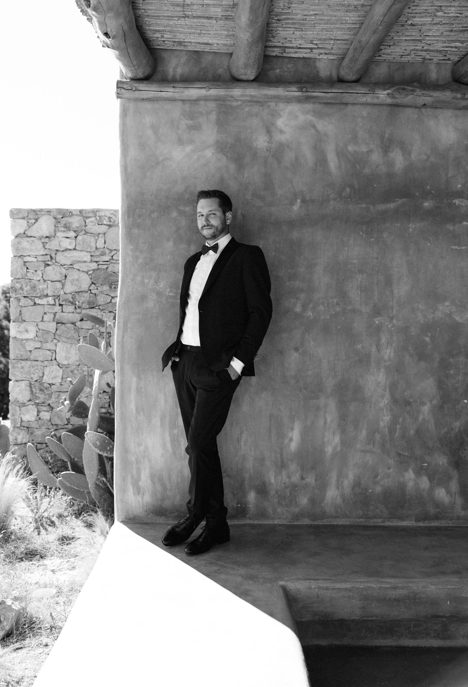 Man in black tuxedo and bow tie leaning against adobe wall in black and white portrait photo.