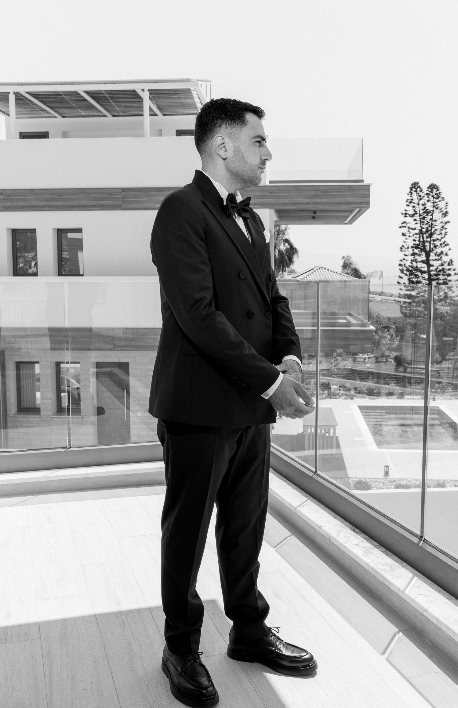 Black and white photograph of a man in a formal suit looking out from a modern building balcony.