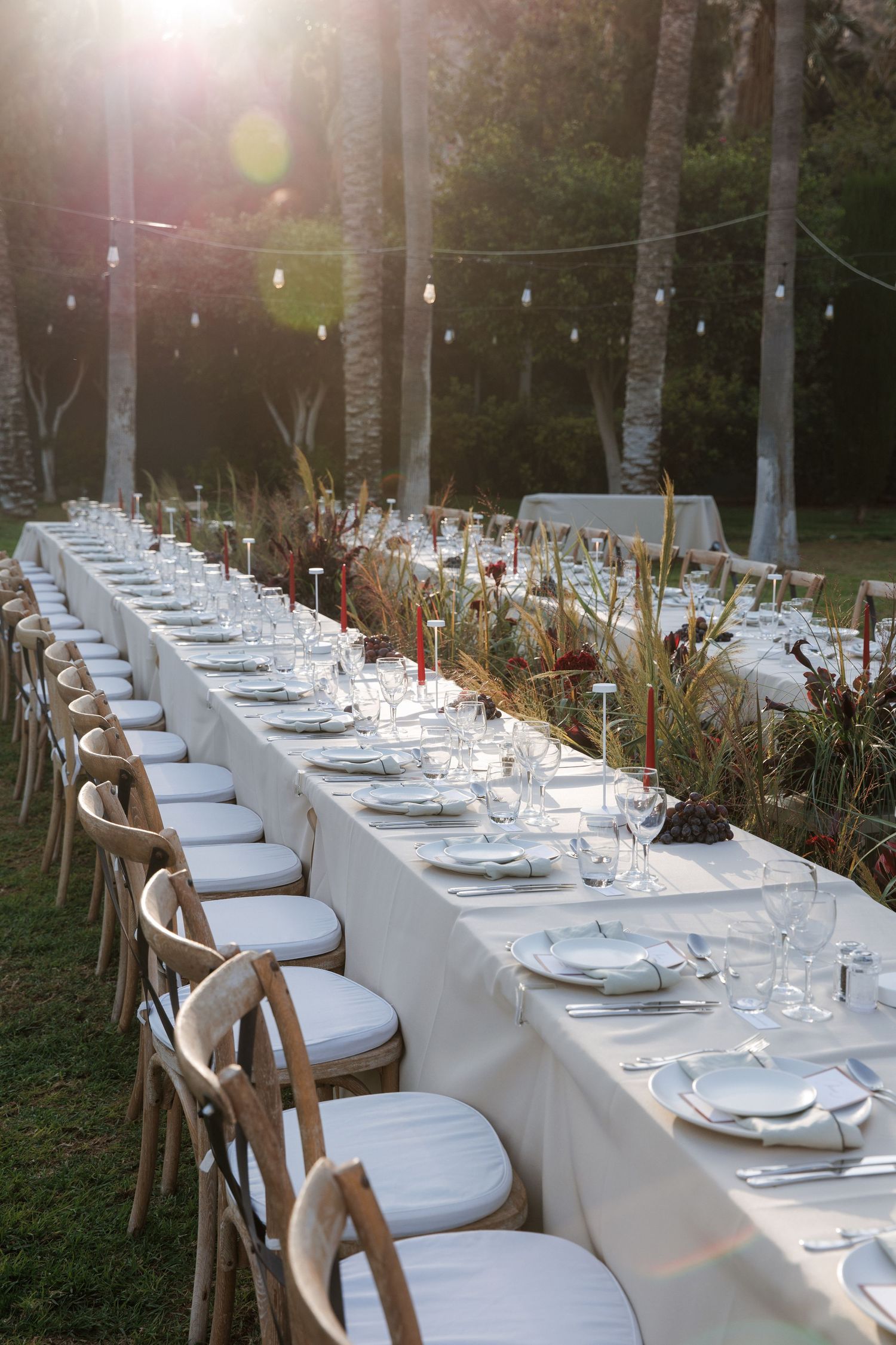 Long outdoor wedding reception table set with white linens, glassware, and floral centerpieces under string lights at sunset.