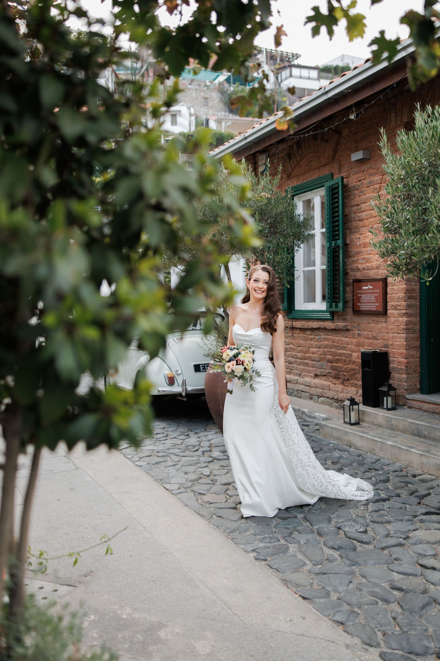 Bride in white gown stands by rustic brick building with green foliage.