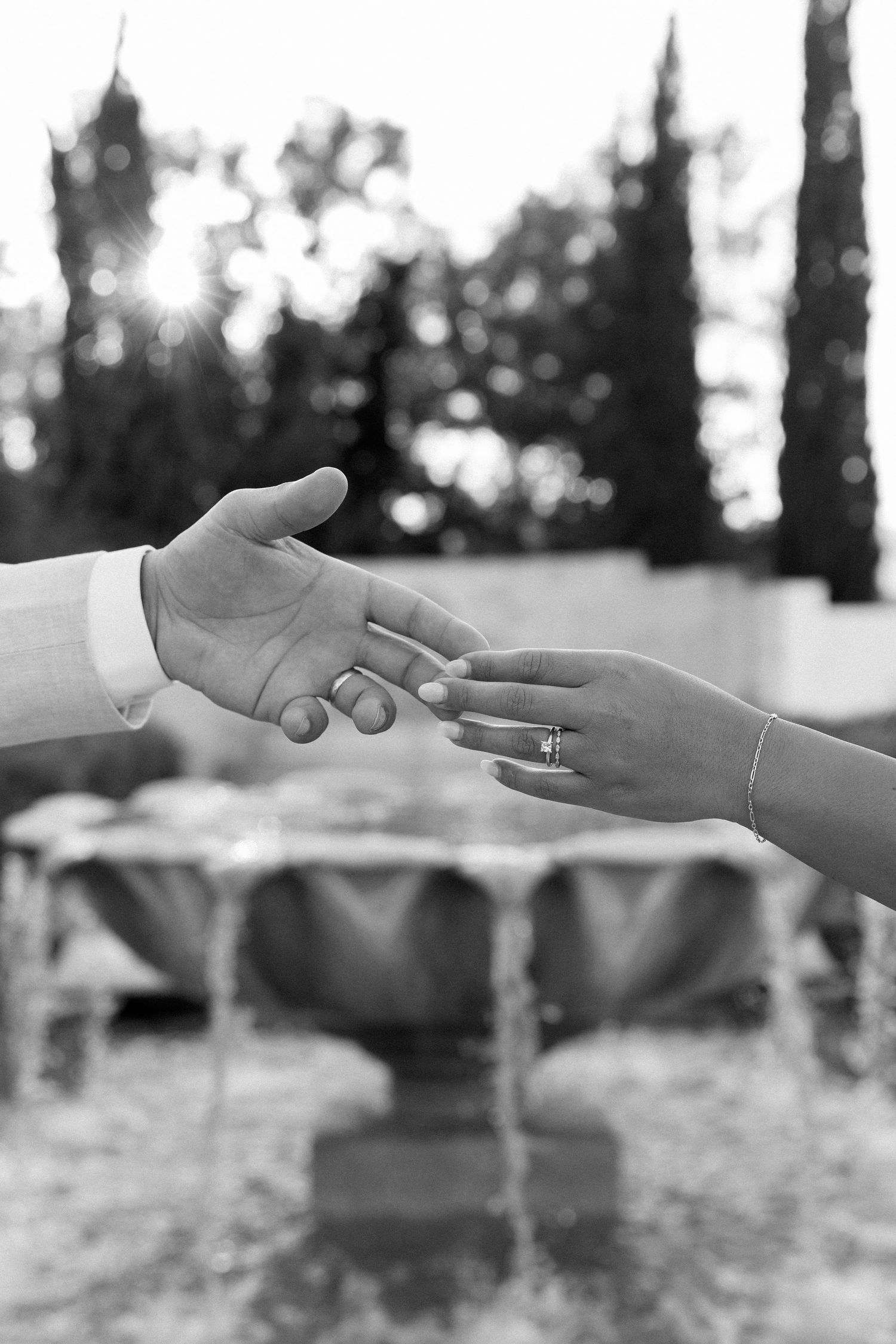 Black and white photo of bride and groom reaching hands toward each other, wedding rings visible, fountain in background.