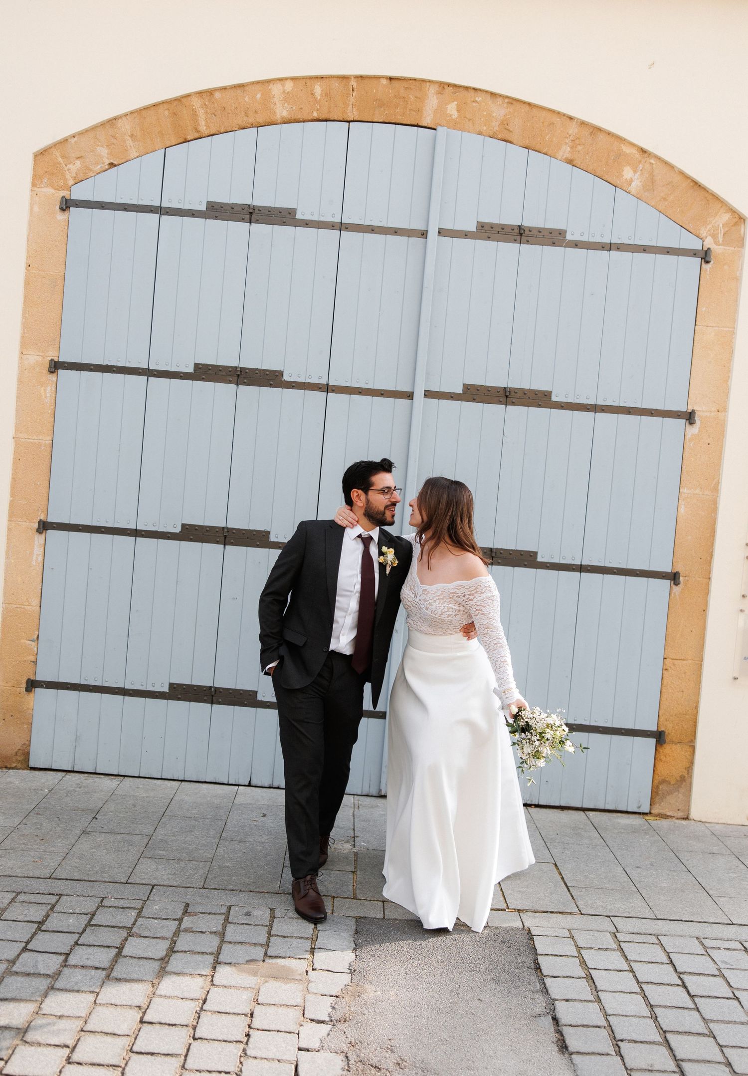 Bride and groom stand facing each other before large light blue arched doors, bride holding white bouquet.