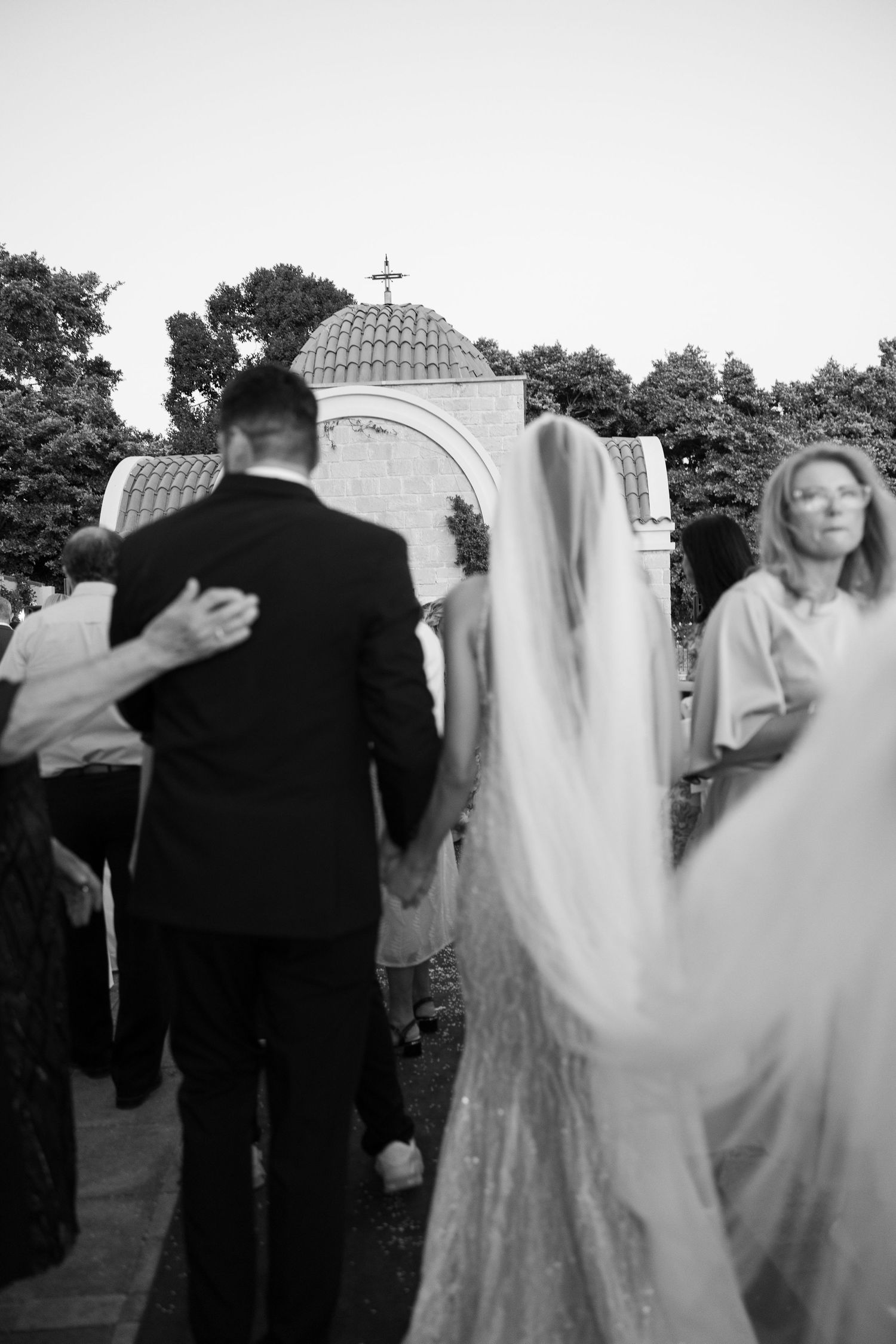 Black and white photo of bride and groom walking toward a chapel with a cross, wedding veil flowing gracefully.