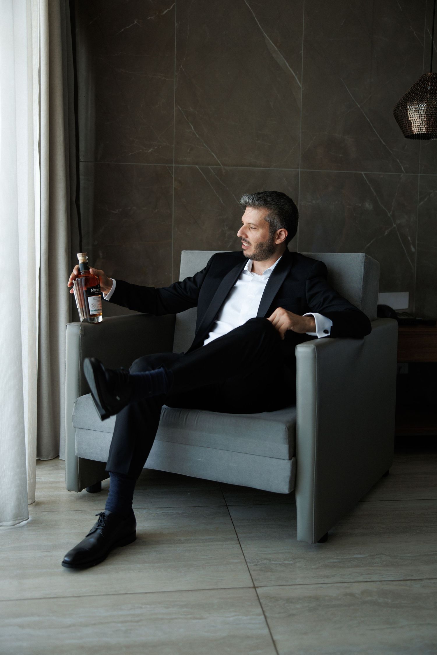 A sophisticated man in a black suit sits in a gray armchair, holding a whiskey bottle while gazing toward a window.