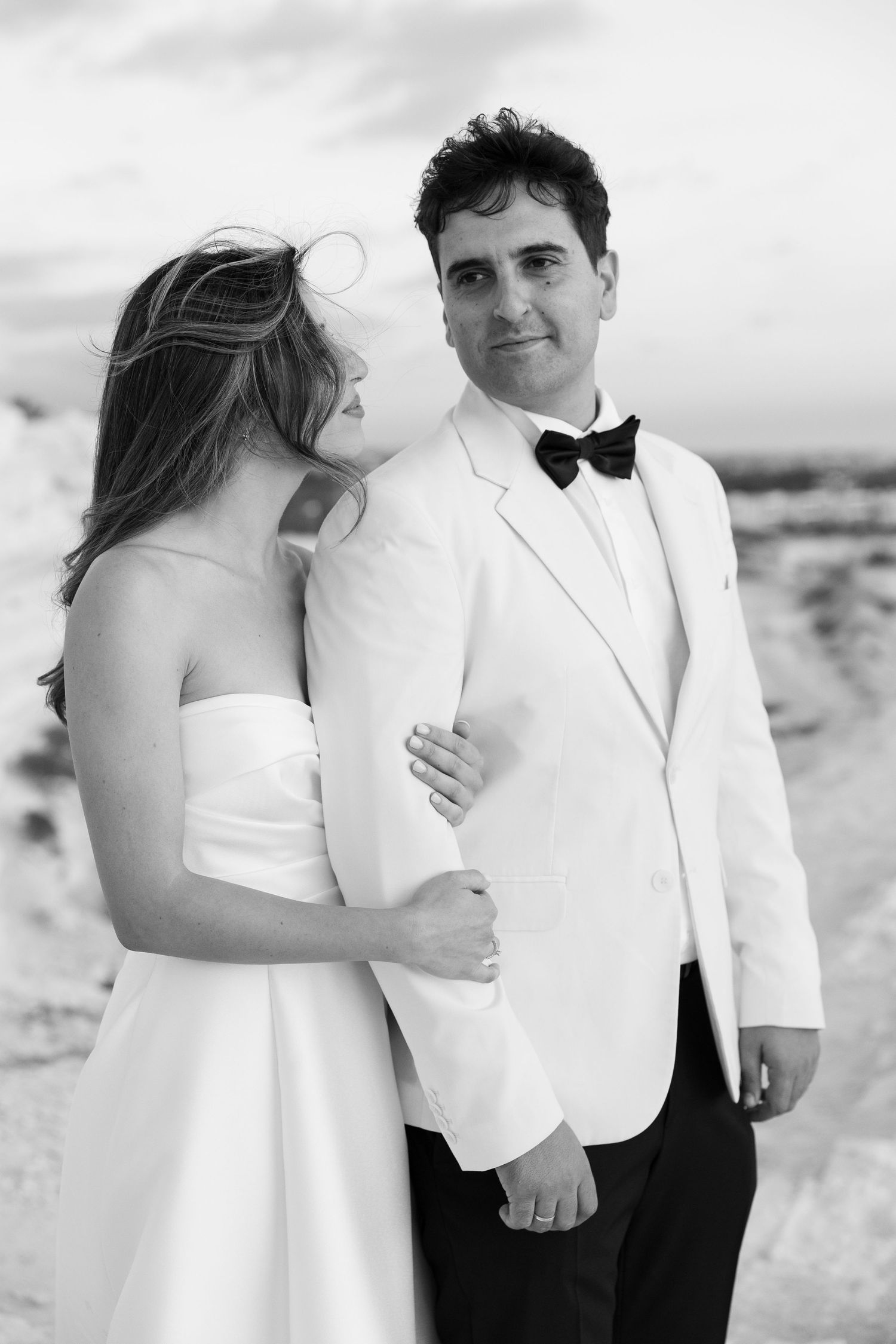 Bride and groom pose together outdoors, groom in white tuxedo with bow tie, elegant black and white portrait.