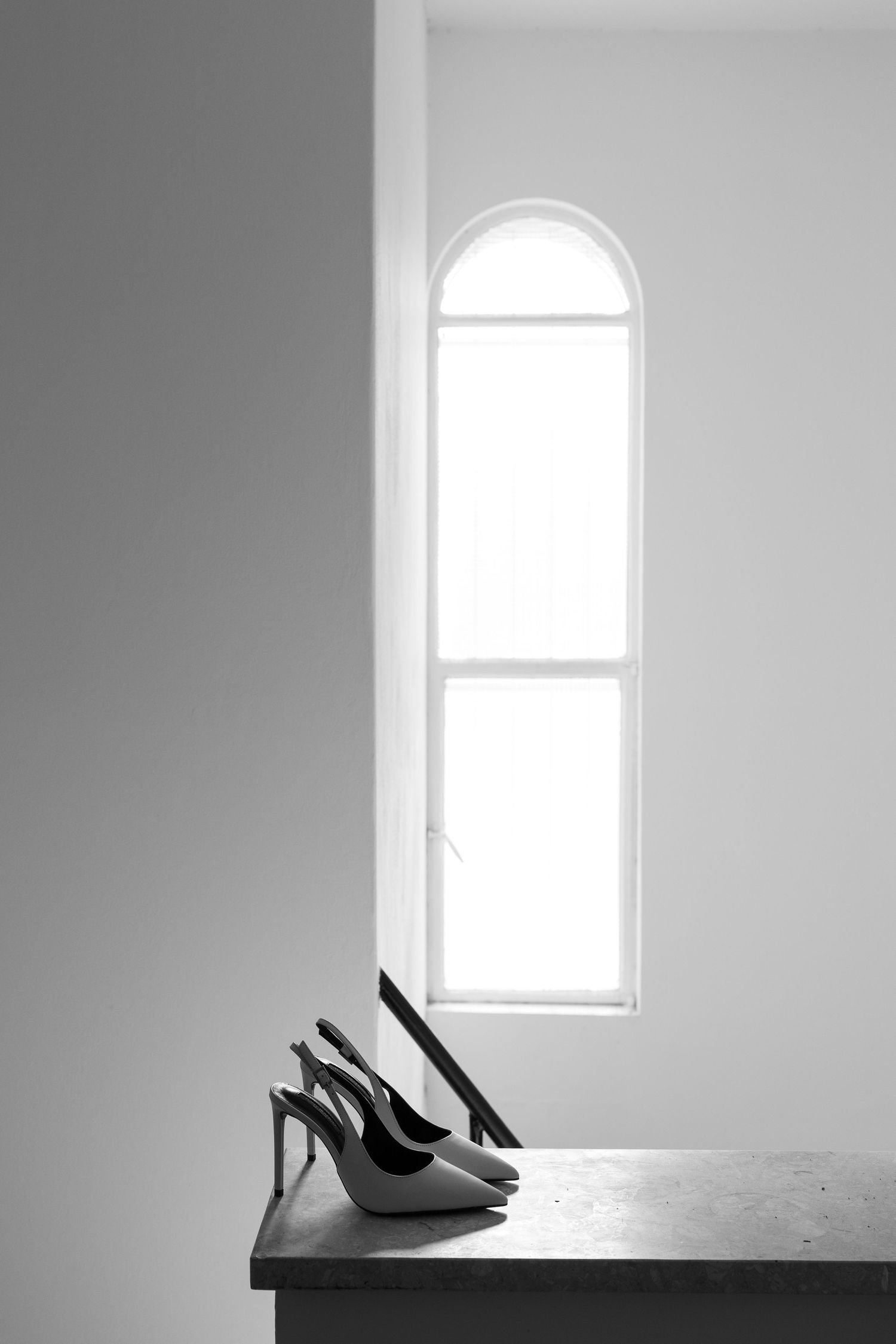 Black and white photo of elegant high heel shoes on a table beside an arched window in a minimalist room.