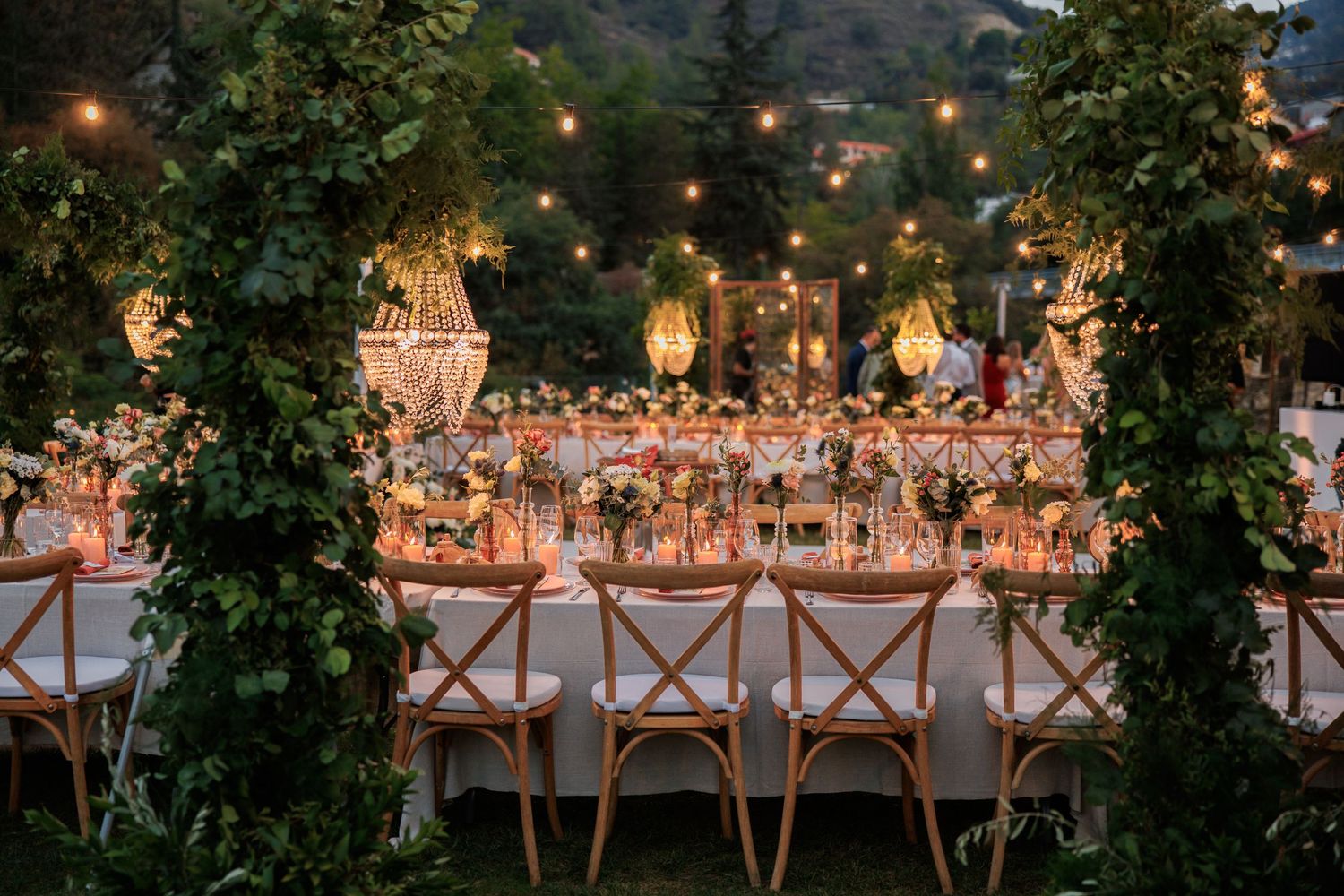Luxurious outdoor dinner setting with hanging chandeliers and cross-back chairs under string lights.