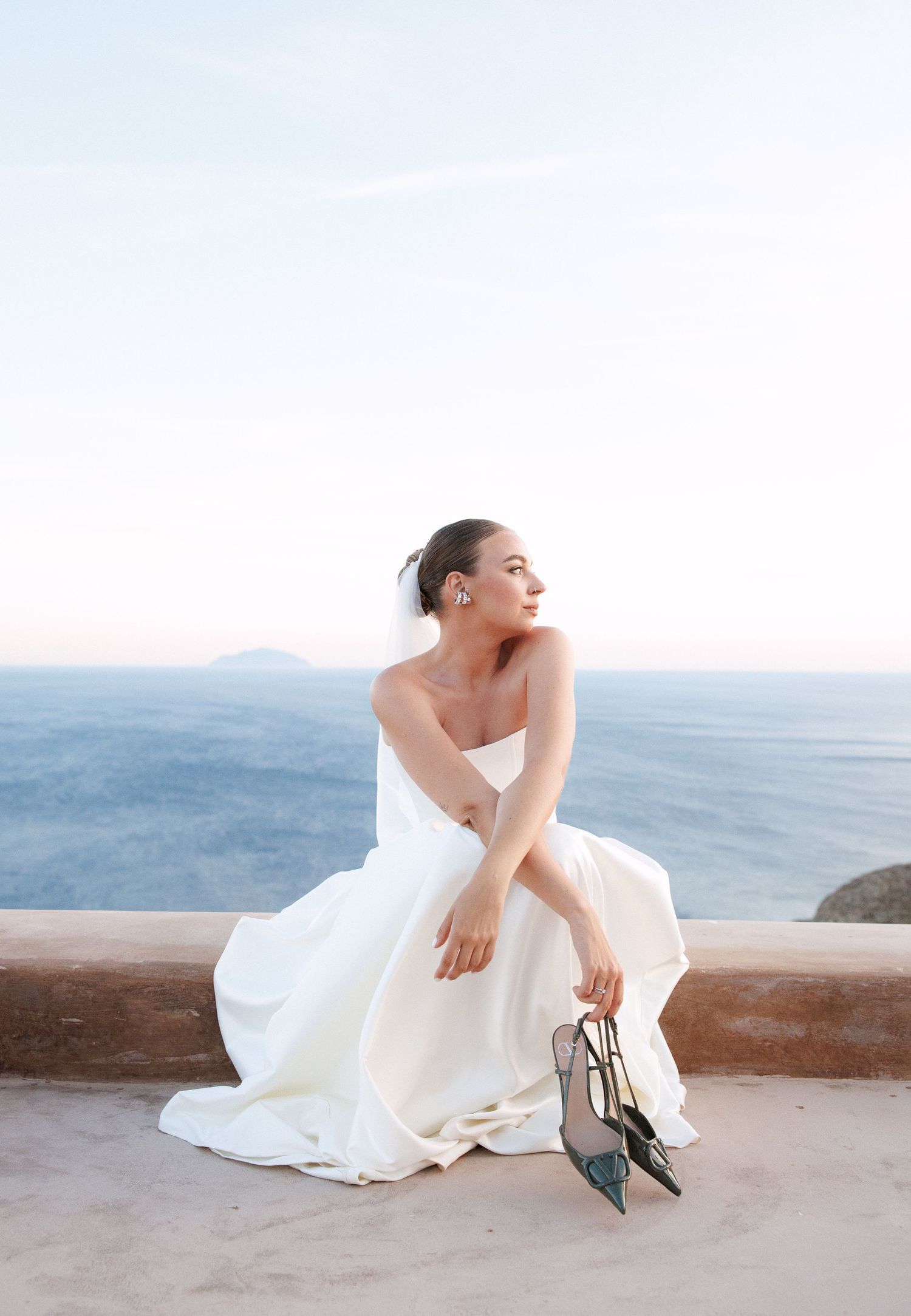 Bride in white strapless gown holds heels while seated on stone wall with ocean view at sunset.