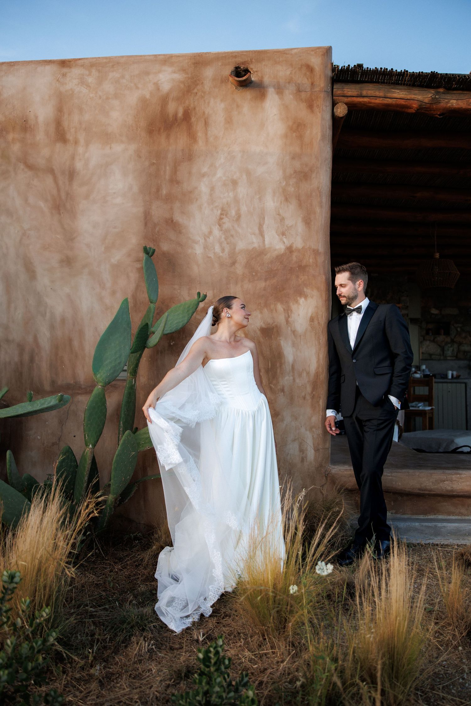 Bride in white strapless gown and groom in black tuxedo stand near adobe wall with cactus at outdoor wedding.
