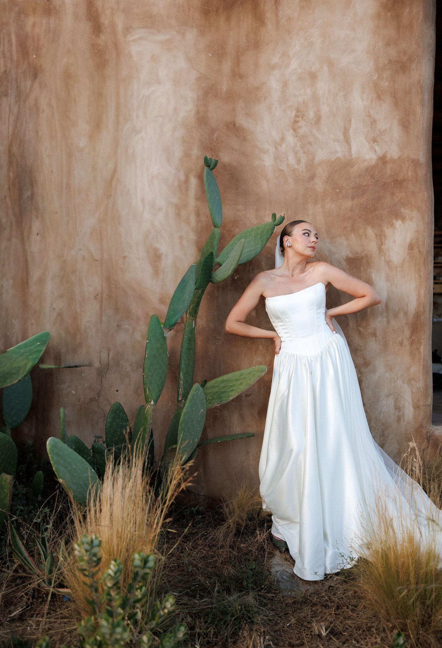 Bride in strapless white wedding gown with train poses by adobe wall with prickly pear cactus in desert setting.