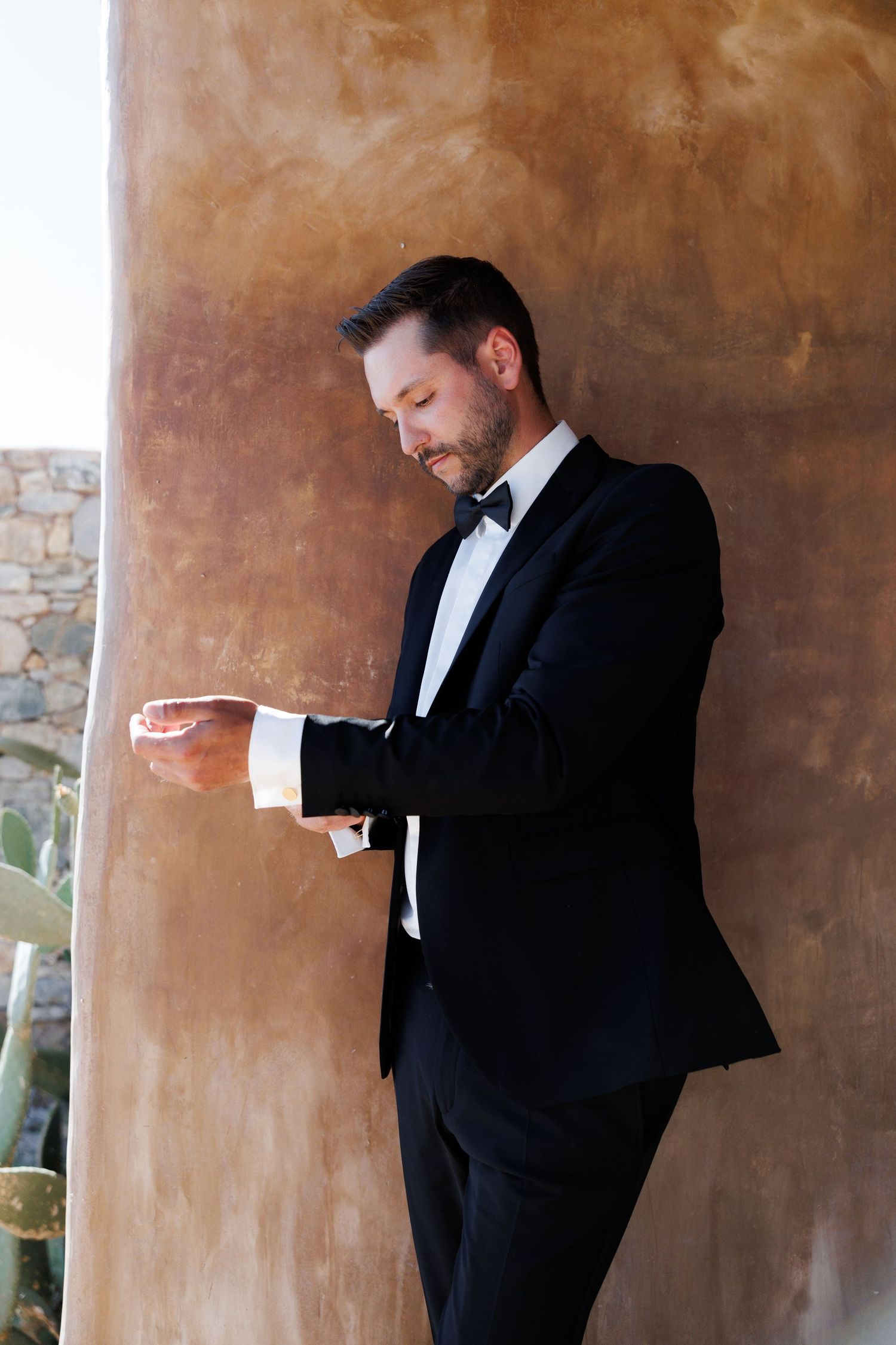 Groom in black tuxedo with bow tie adjusting cufflinks against a rustic terracotta wall.