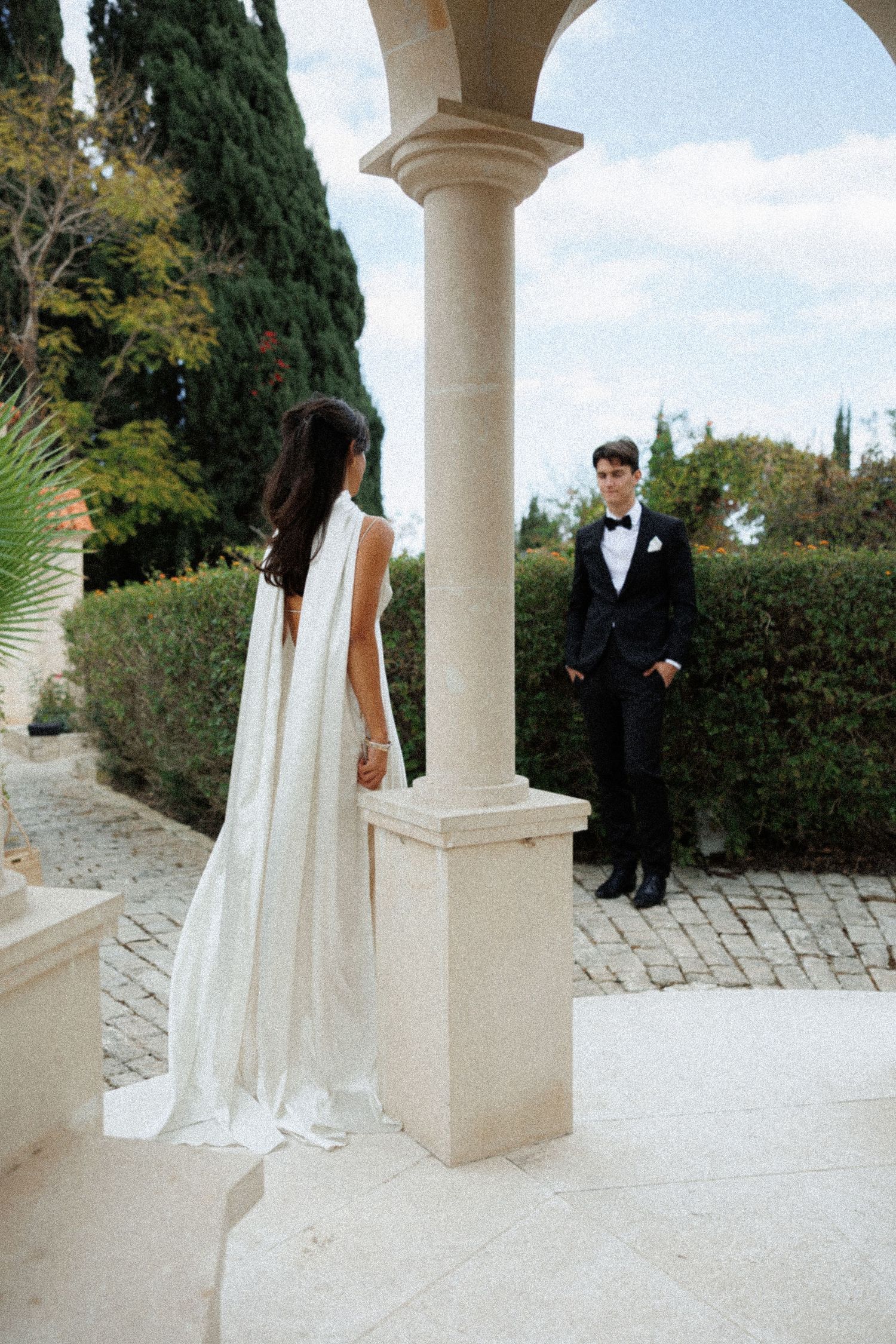A couple in wedding attire stands on an elegant stone patio with classical columns and cypress trees in the background.
