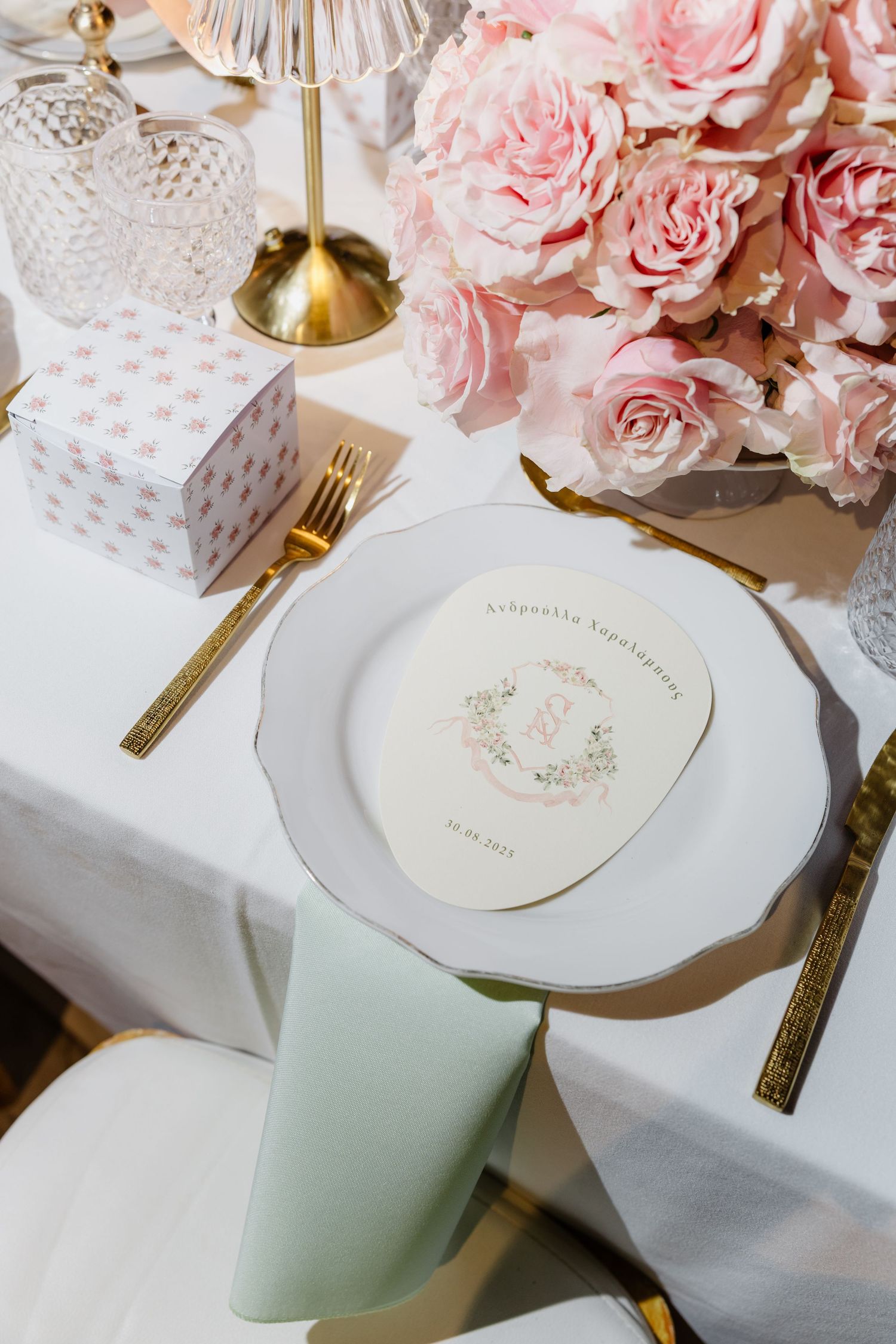 Elegant table setting with pink roses, crystal glassware, gold cutlery, and decorative plates on white tablecloth.