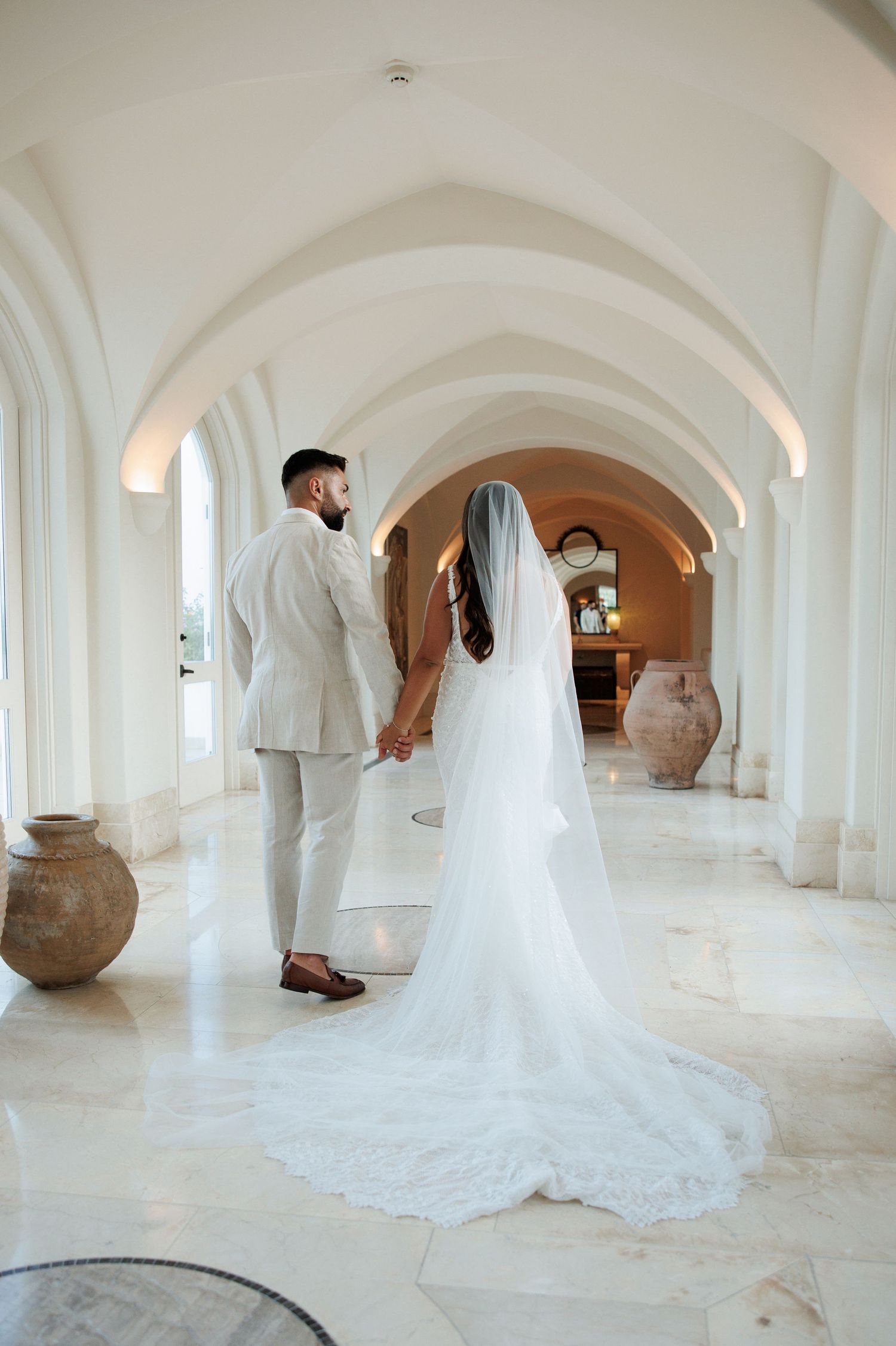 Bride and groom holding hands walking through elegant white arched hallway, bride wearing long cathedral veil.