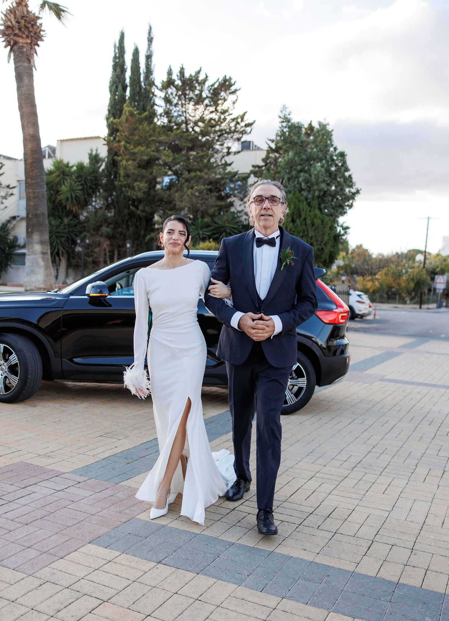 Elegant bride in white gown with slit walks alongside man in navy tuxedo on paved plaza near black SUV.