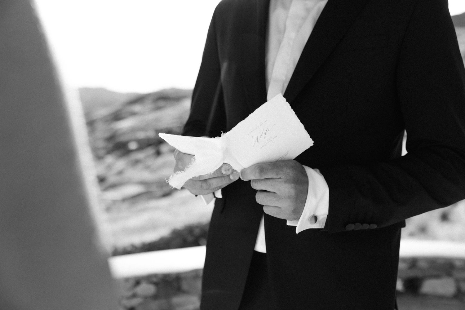 Groom in black suit reading wedding vows from paper during outdoor ceremony in black and white photo.