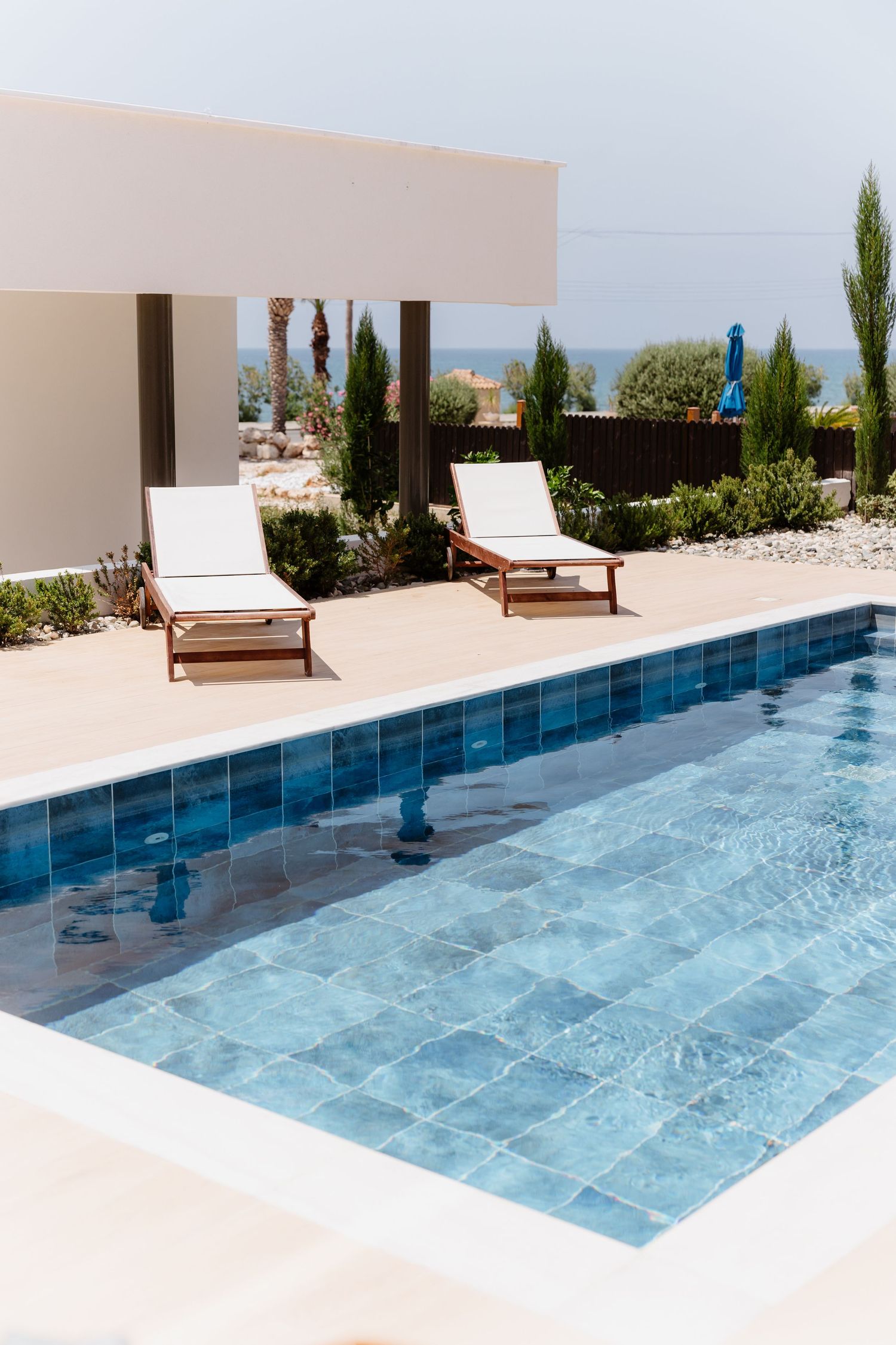 Modern pool terrace with two white loungers overlooking the ocean, featuring tiled blue water and cypress trees in garden.