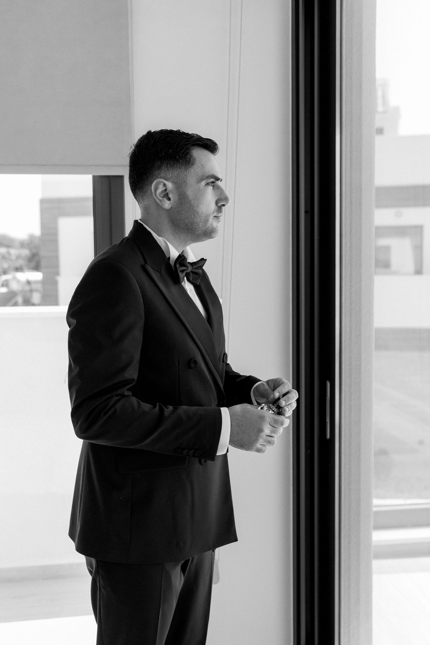 A person in a dark suit stands contemplatively by large windows in a black and white photograph.