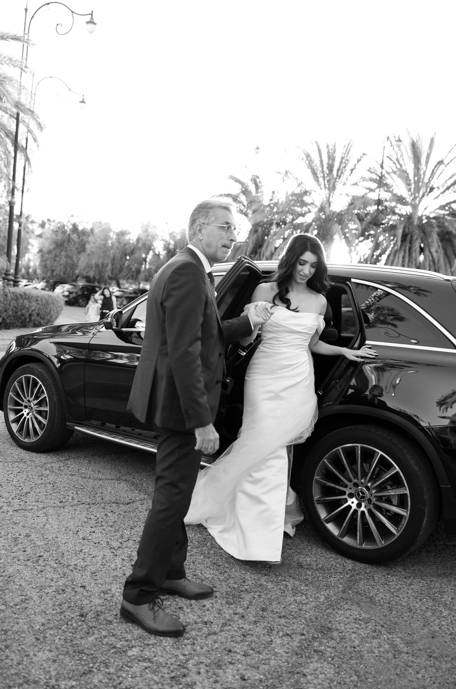 A bride in a white gown steps out of a luxury black SUV assisted by a man in a suit, in a black and white photo.