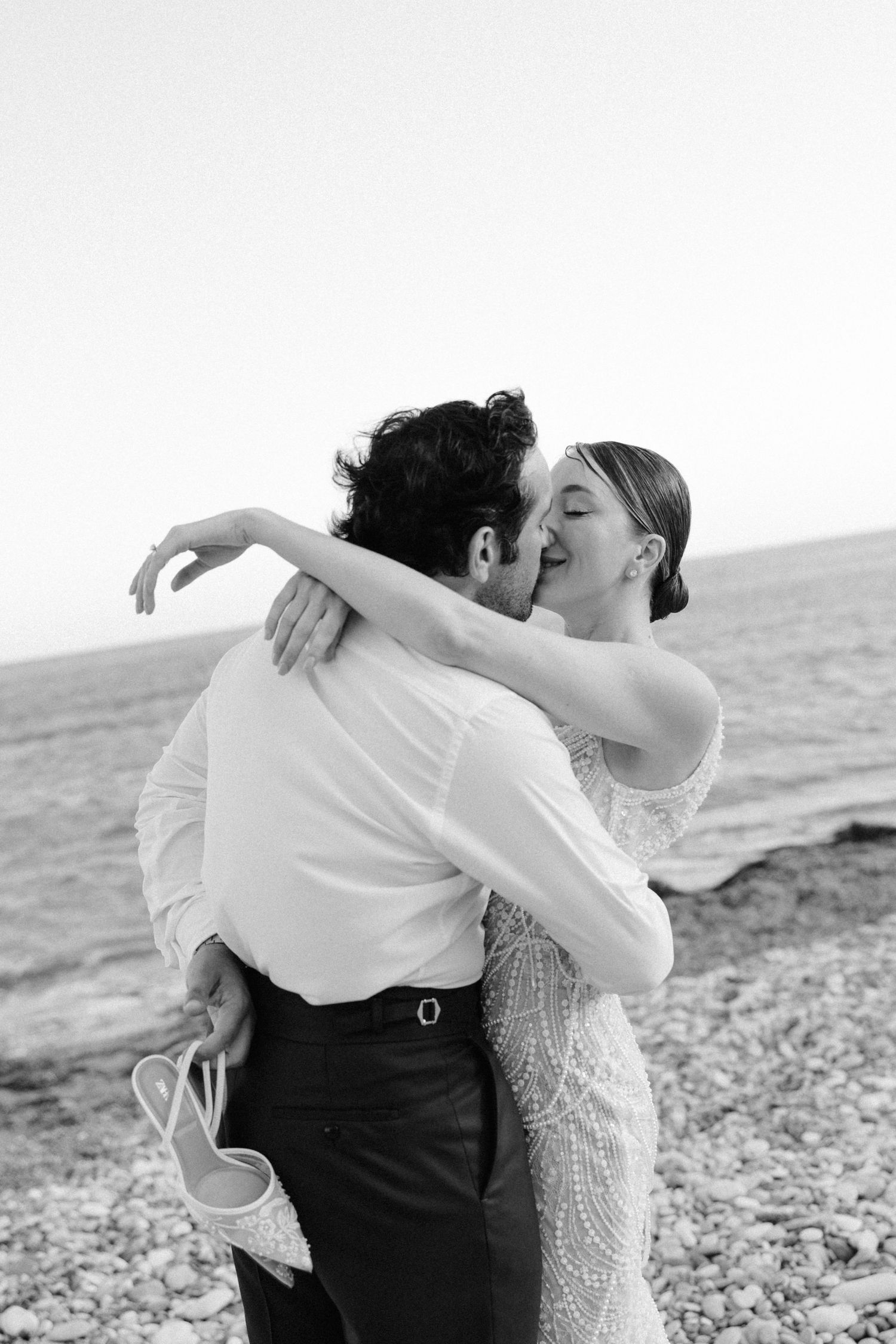 Black and white photo of a couple embracing and kissing on a rocky beach, bride in lace gown holding heels.