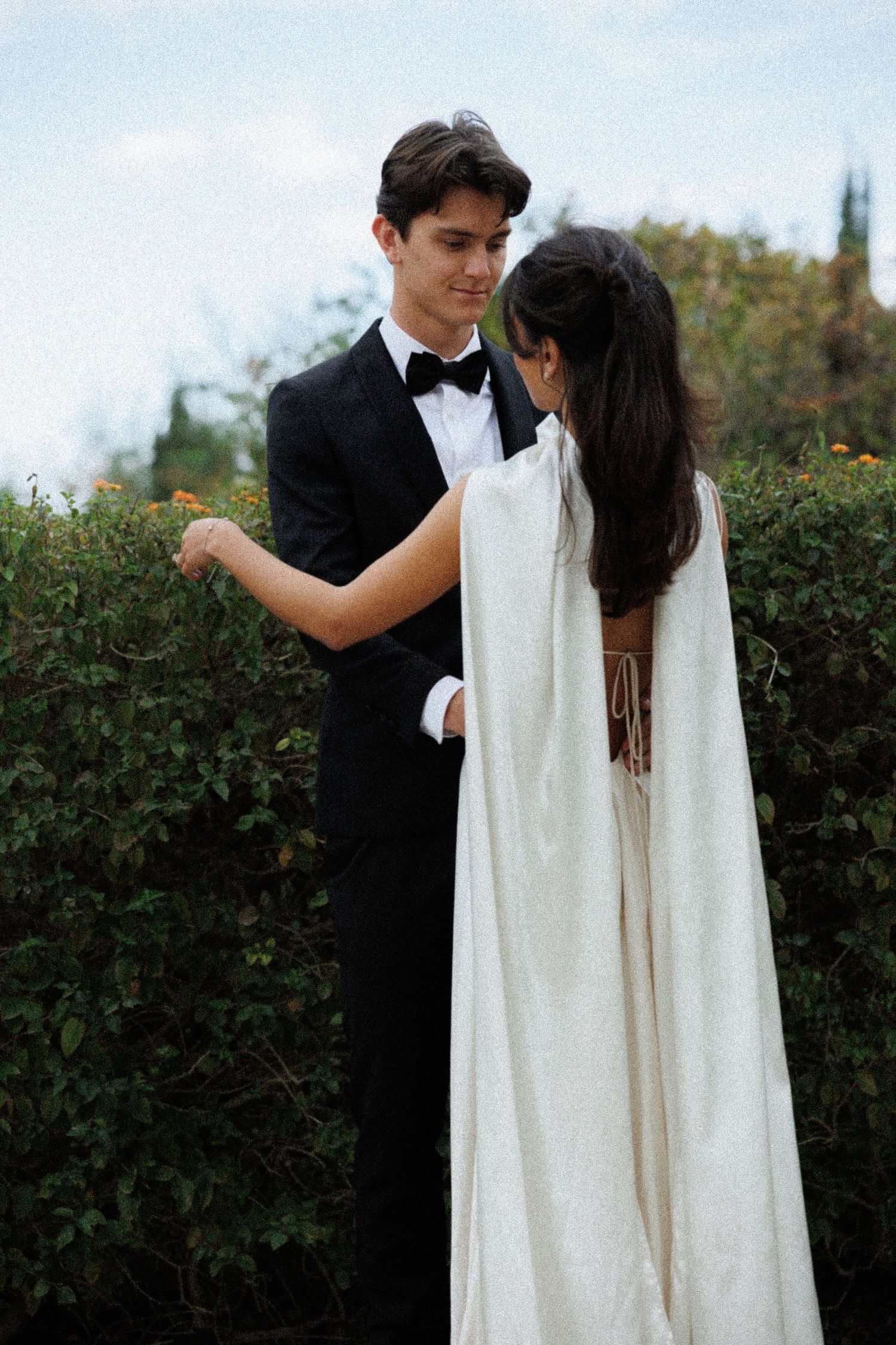 A couple dances together in formal wear surrounded by garden greenery.
