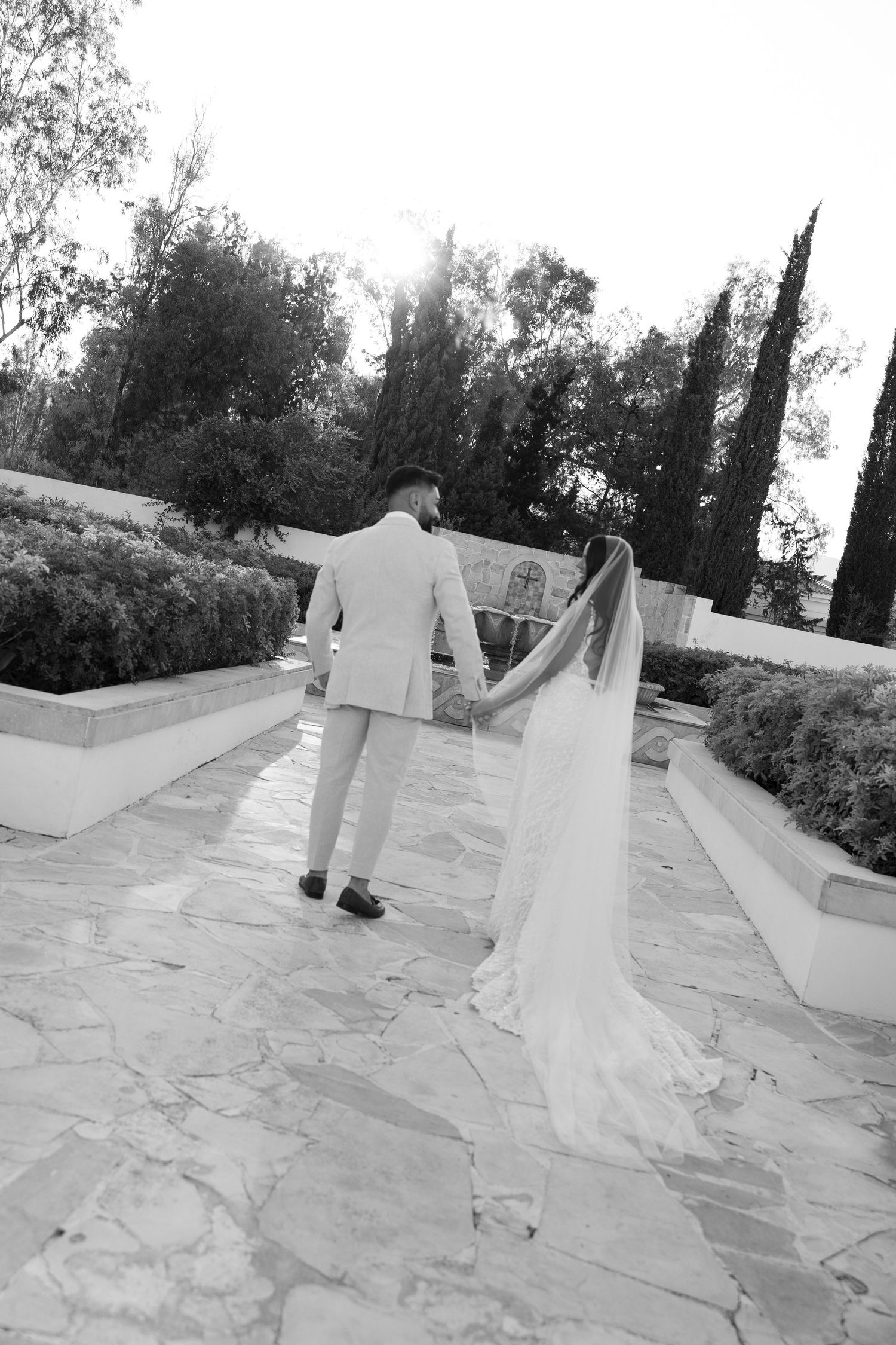 Black and white photo of bride and groom holding hands walking through elegant outdoor garden venue.