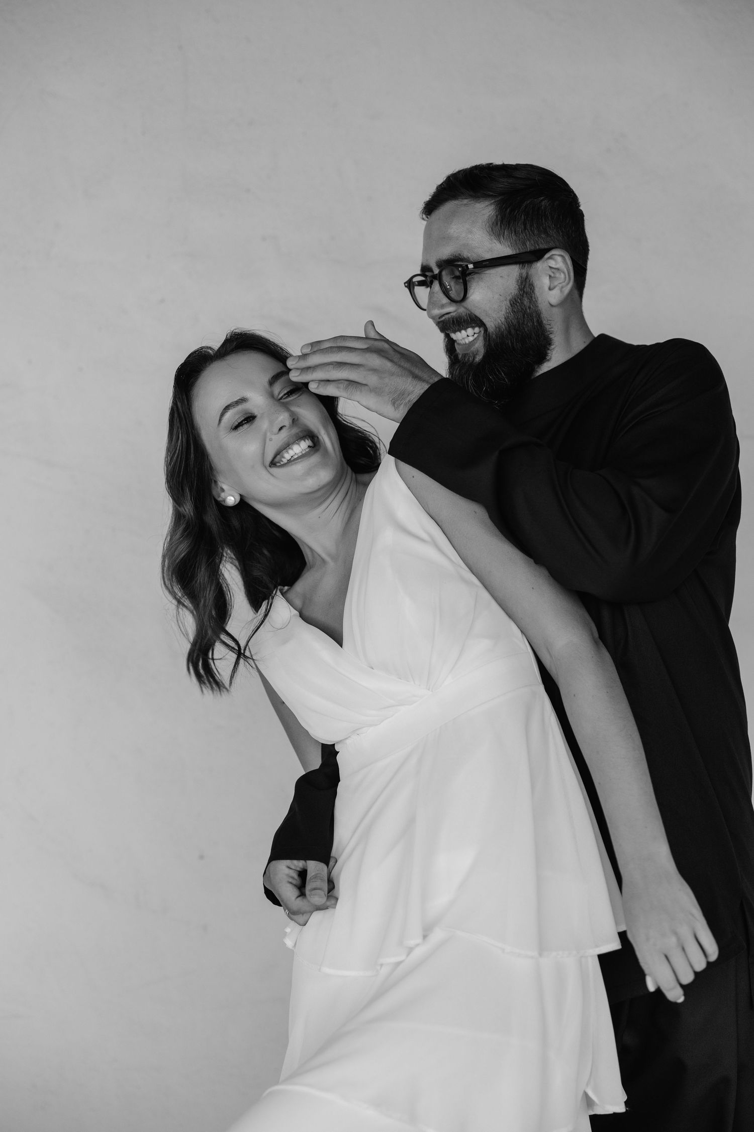 Black and white photo of a joyful couple laughing together against a plain wall, sharing a playful moment.