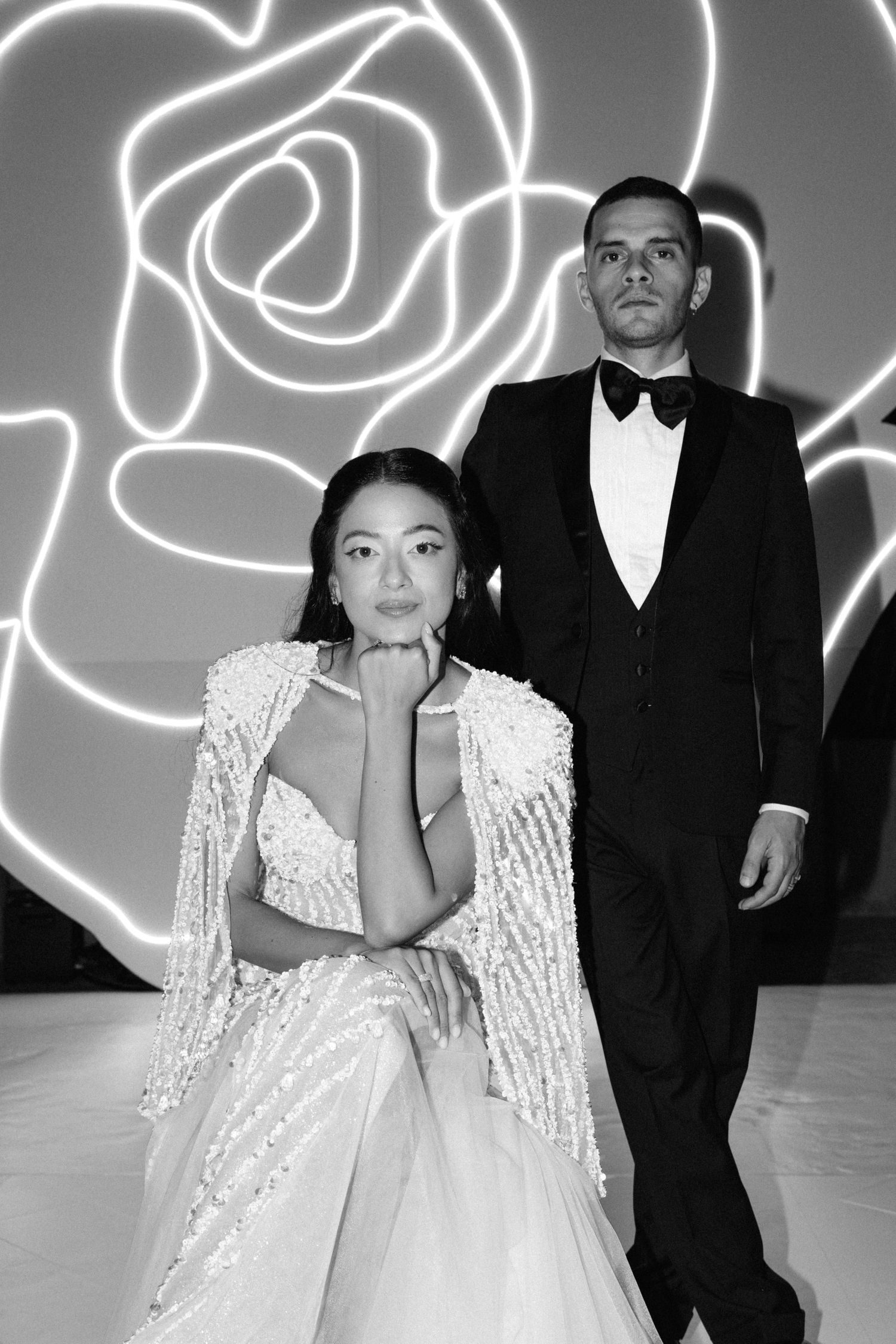 Black and white photo of a couple in formal wear, woman in sequined gown, man in tuxedo, rose neon light backdrop.