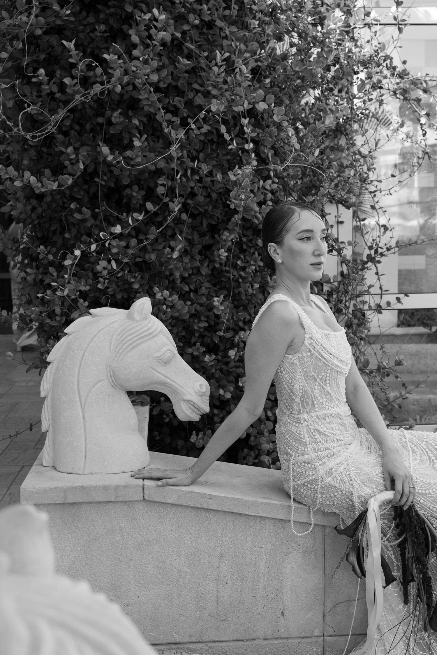 Black and white photo of elegant woman in lace dress posing beside a marble horse sculpture with lush greenery backdrop.