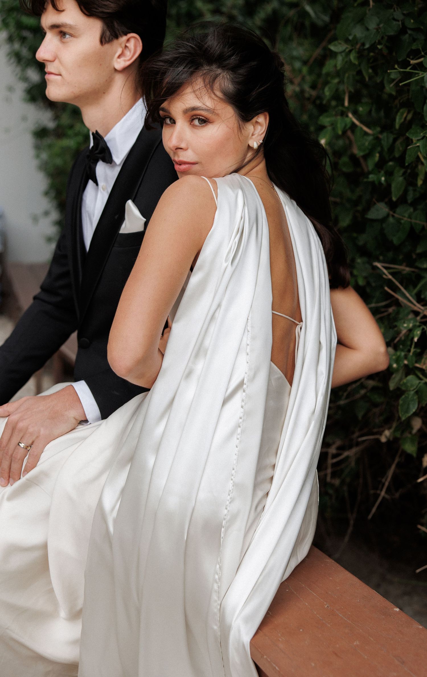 A wedding couple poses together outdoors, with the bride wearing an elegant backless white dress with flowing fabric details.