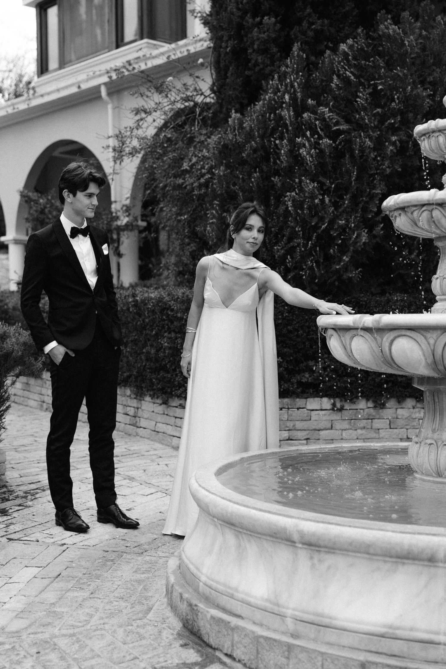 Black and white romantic photo of a couple in formal wear standing by an ornate stone fountain at an elegant venue.