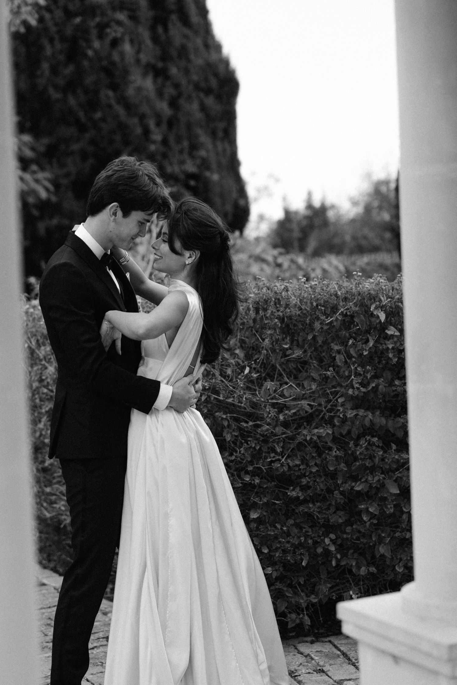 A romantic black and white wedding photo of a couple embracing near a stone column and garden hedge.
