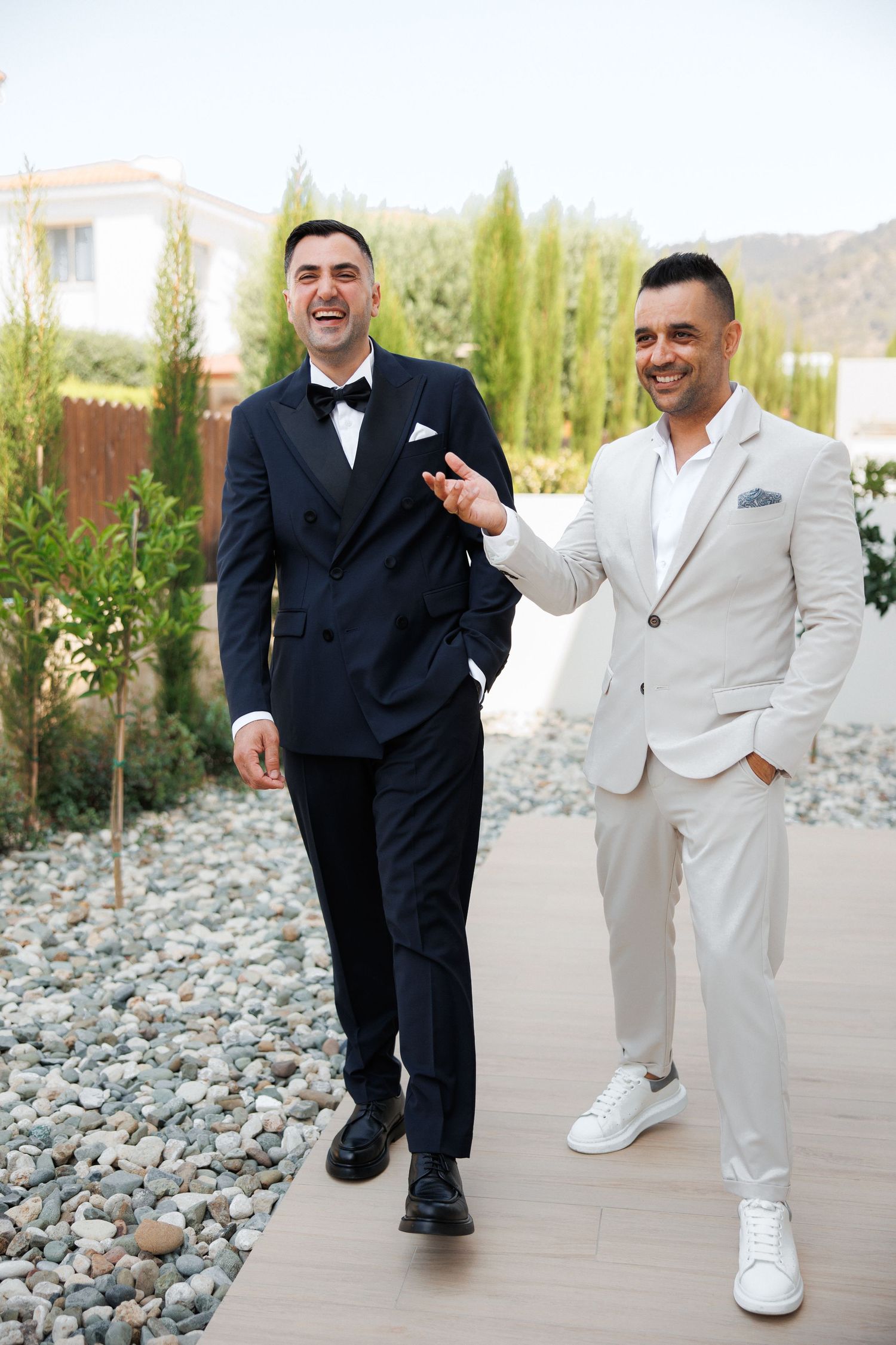 Two formal suit-clad figures pose playfully together on stone path with landscaping visible in background.
