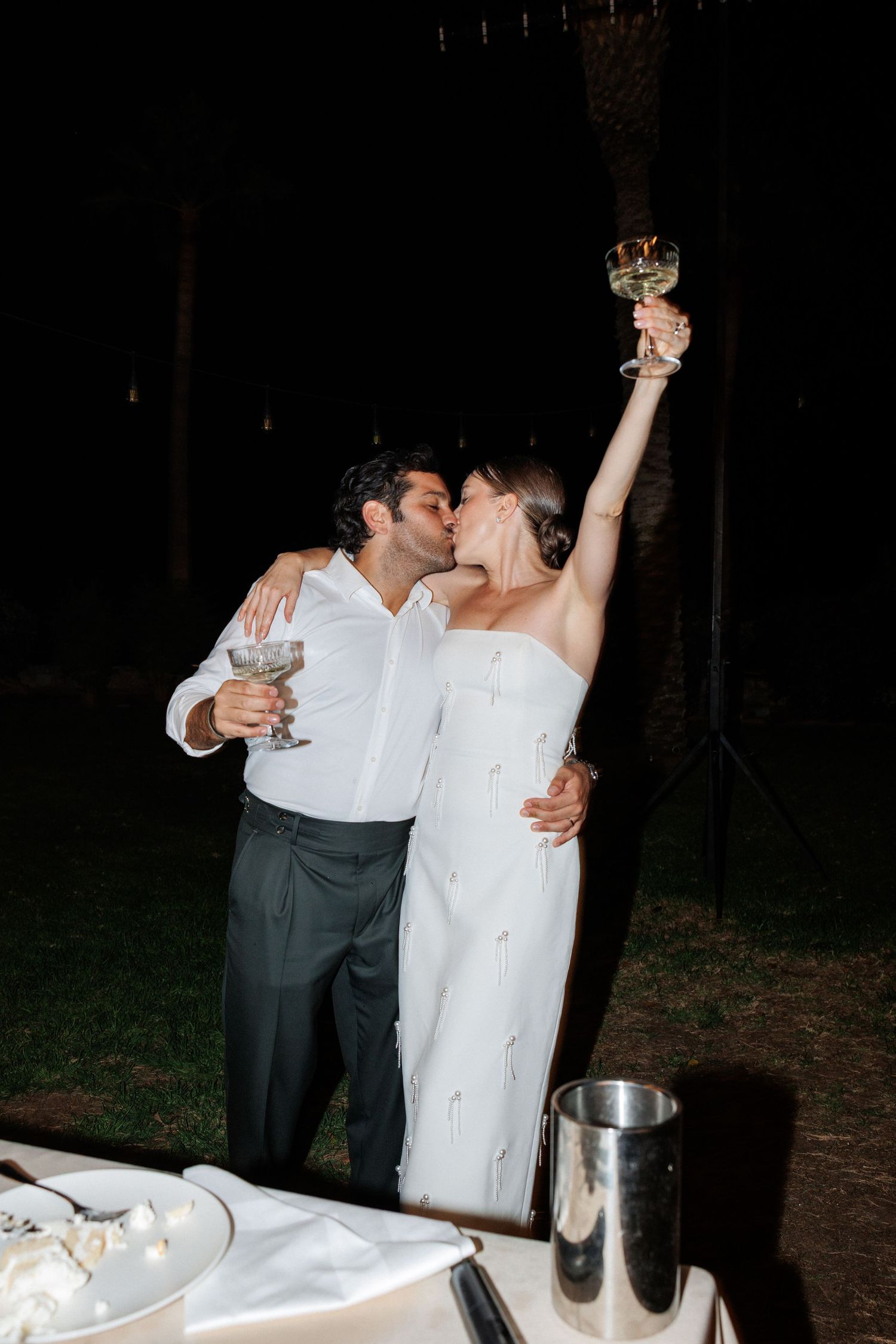 A couple shares a kiss while raising wine glasses in a toast at an outdoor evening wedding reception.