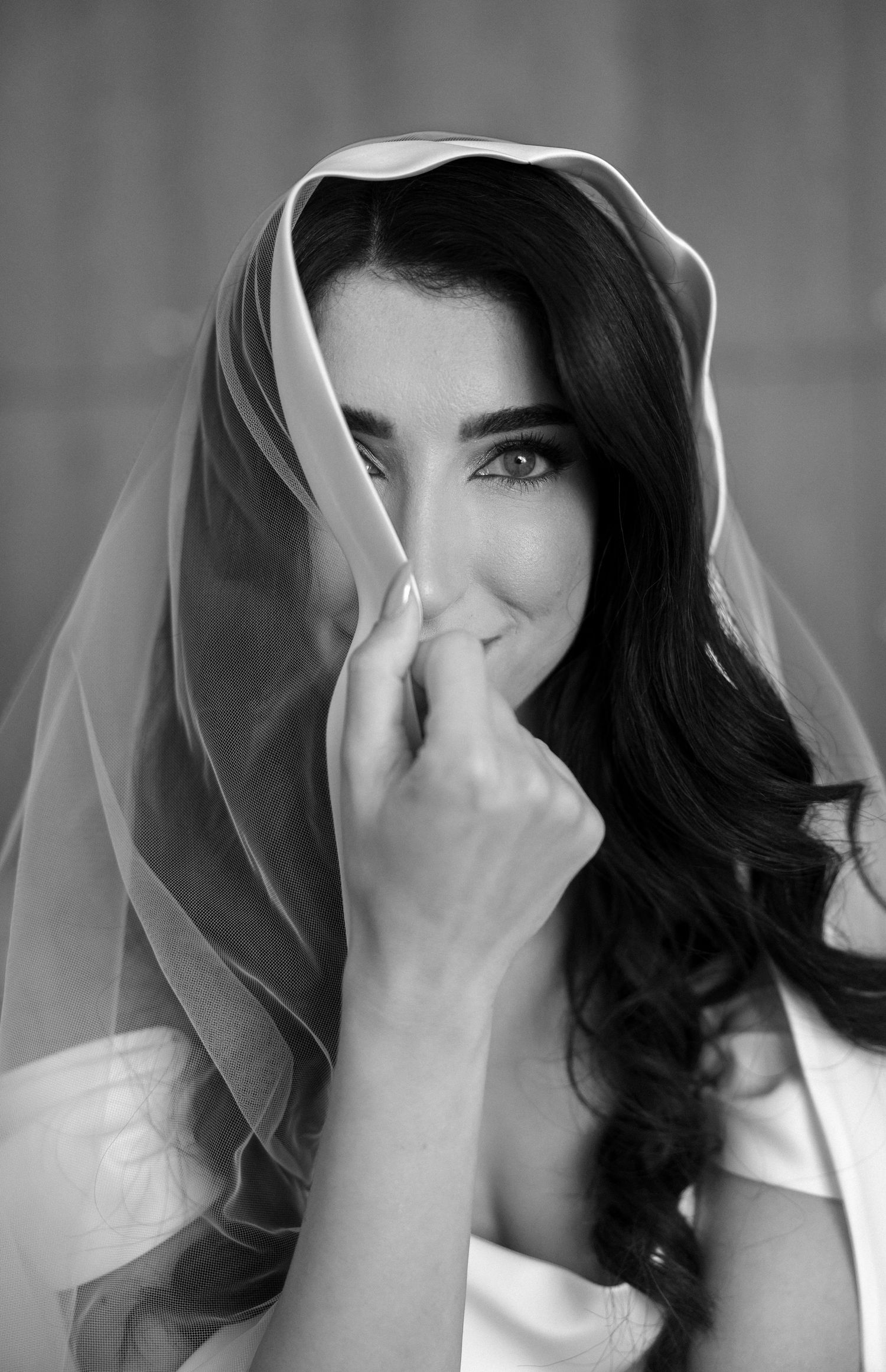 Black and white portrait of a bride holding her veil over her face, with long dark hair and striking eyes.