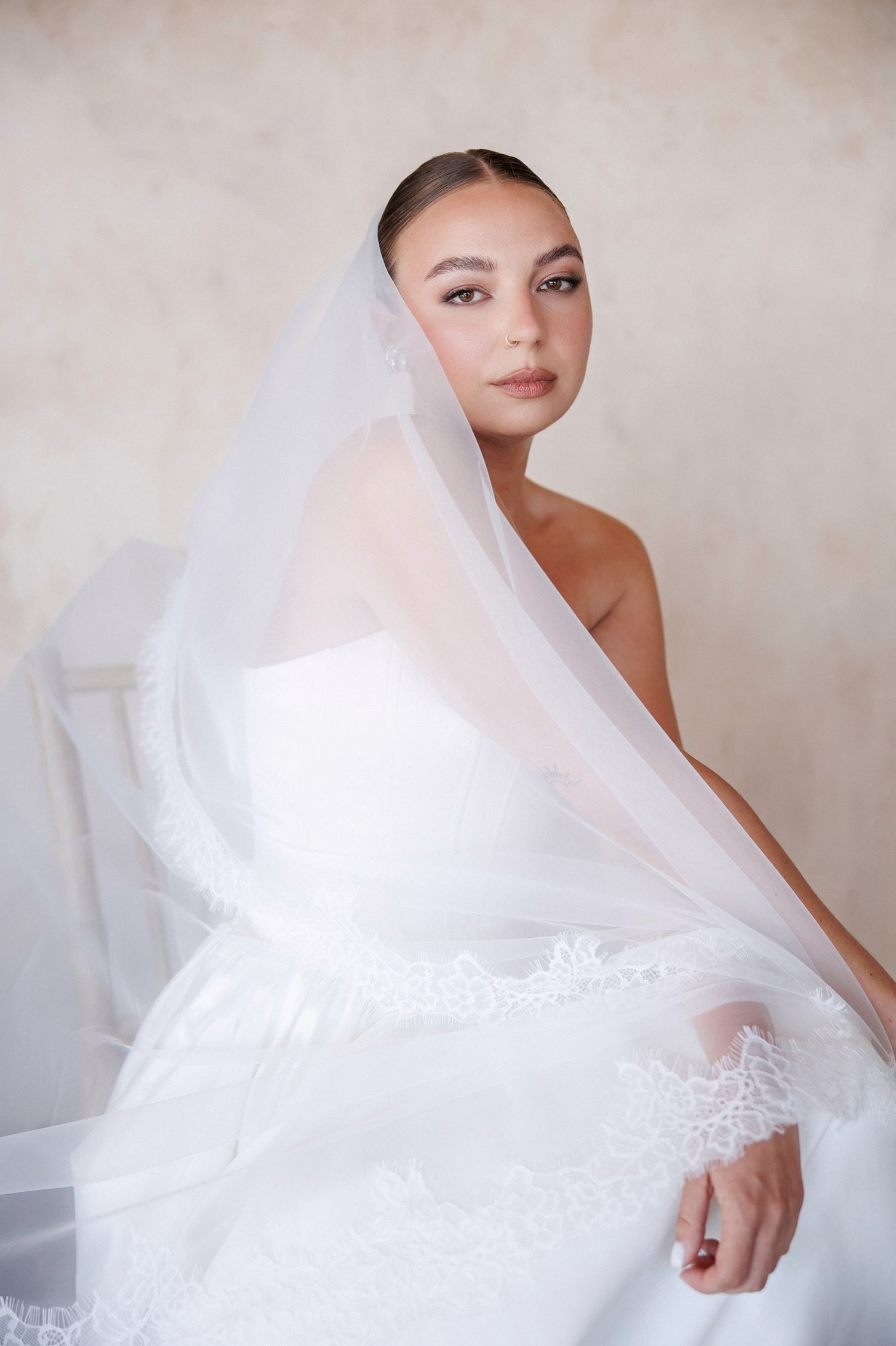 Elegant bride in white lace gown and veil seated gracefully in a softly lit studio setting.