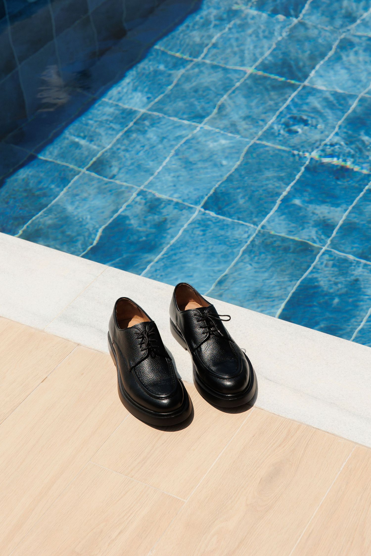 A pair of black leather oxford dress shoes sits on a beige surface next to blue tiled pool water.